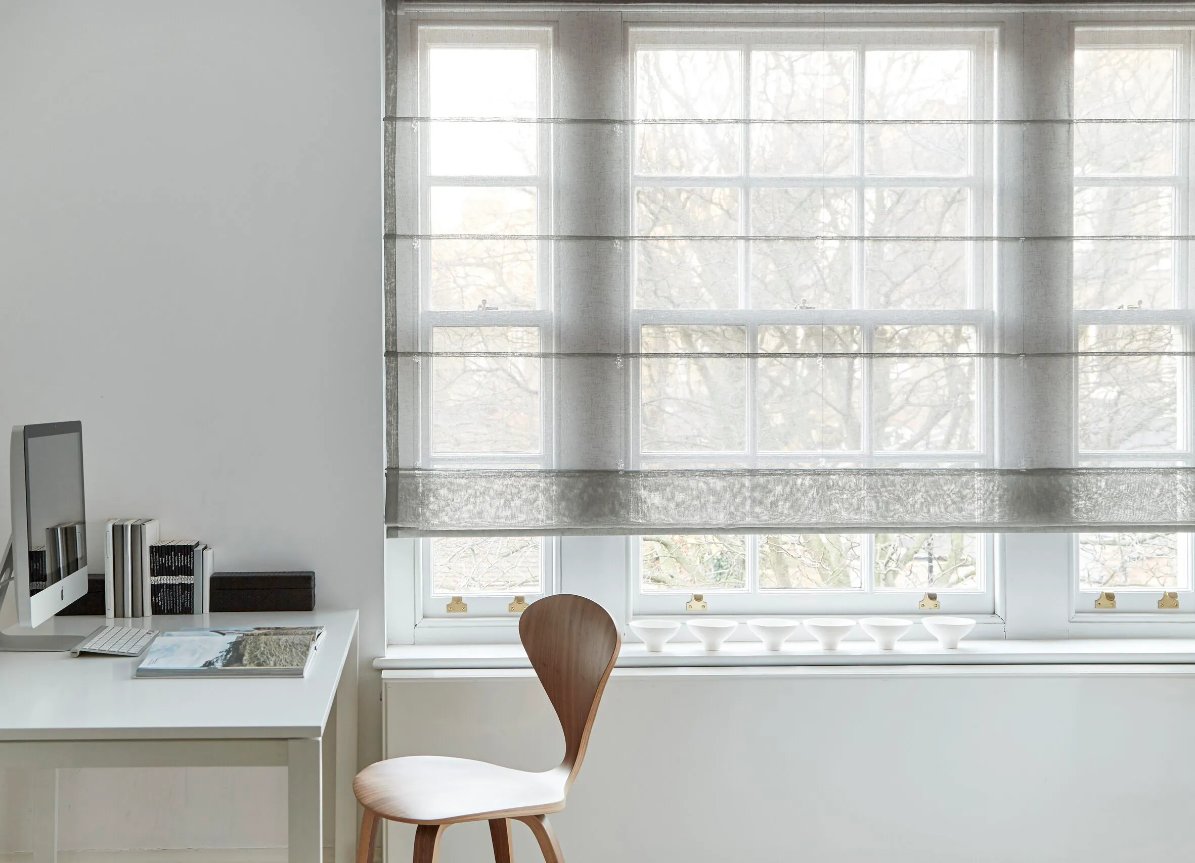 Sheer grey Roman blind softens light at a large multi-pane window beside a minimalist desk and wooden chair.