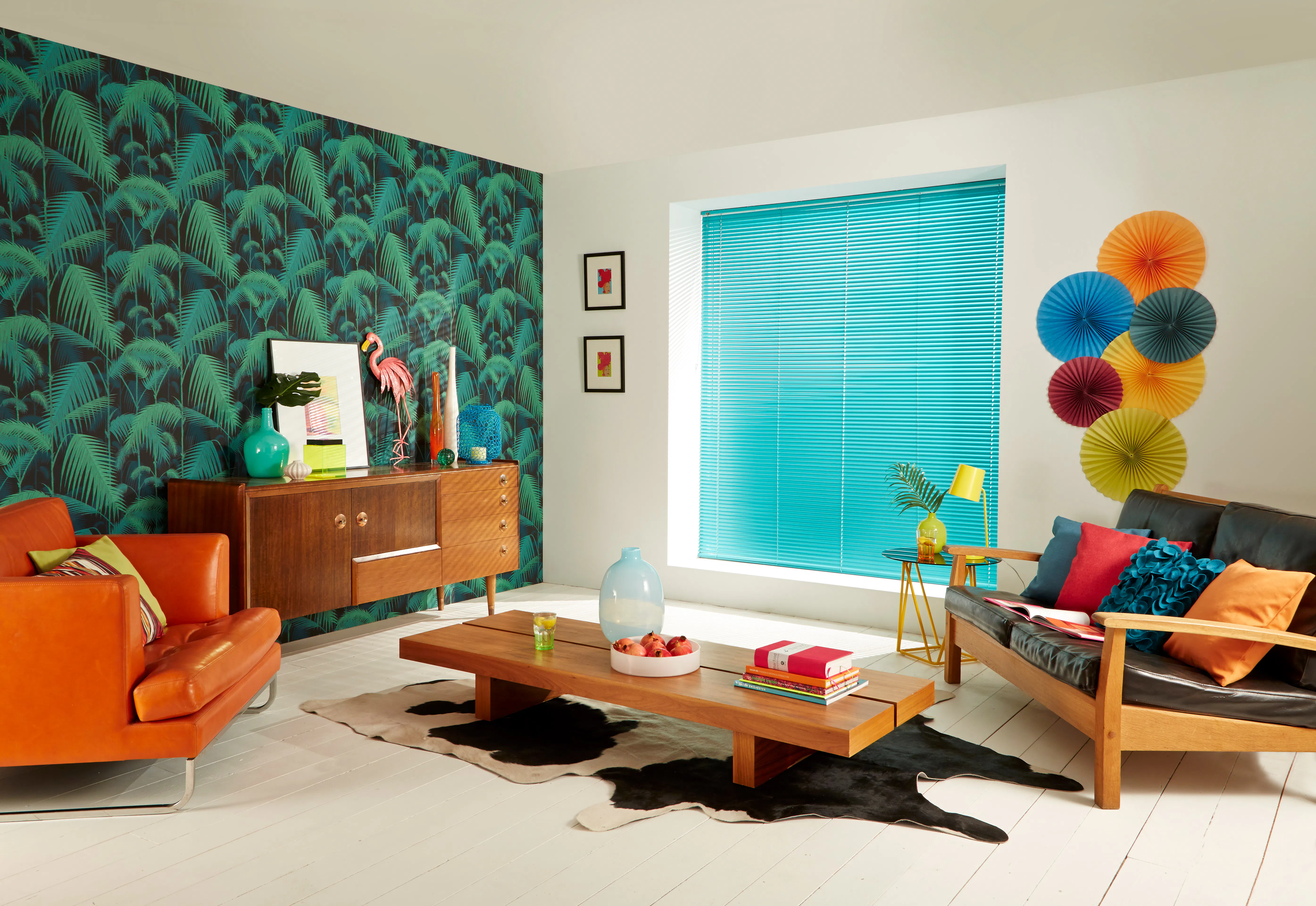 A retro living room with a tropical palm print wallpaper, turquoise Venetian blind, orange leather armchair and a cowhide rug.