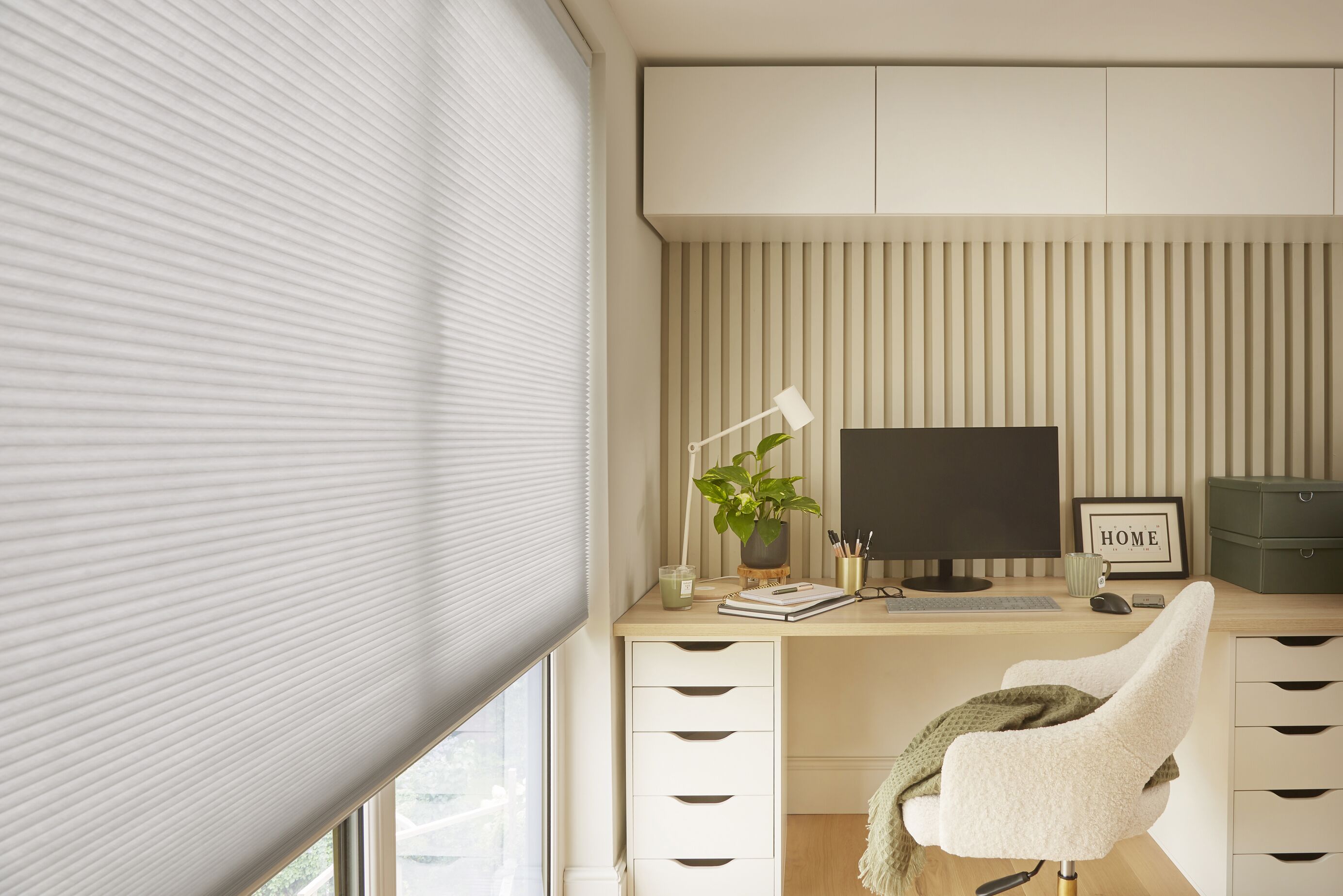 Close-up of a pale grey honeycomb blind beside a fitted desk with monitor, drawers and a cream sherpa office chair.