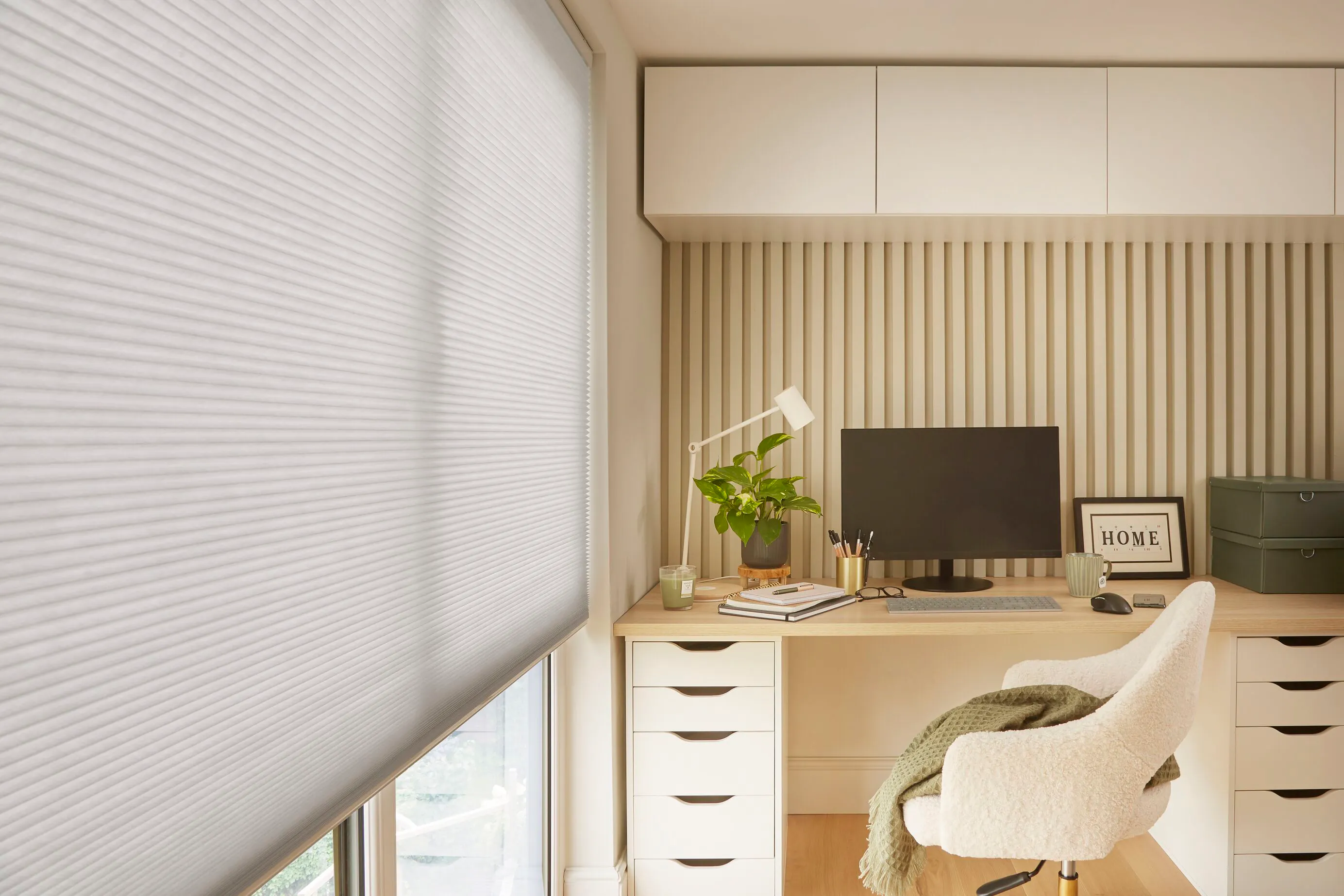 Close-up of a pale grey honeycomb blind beside a fitted desk with monitor, drawers and a cream sherpa office chair.