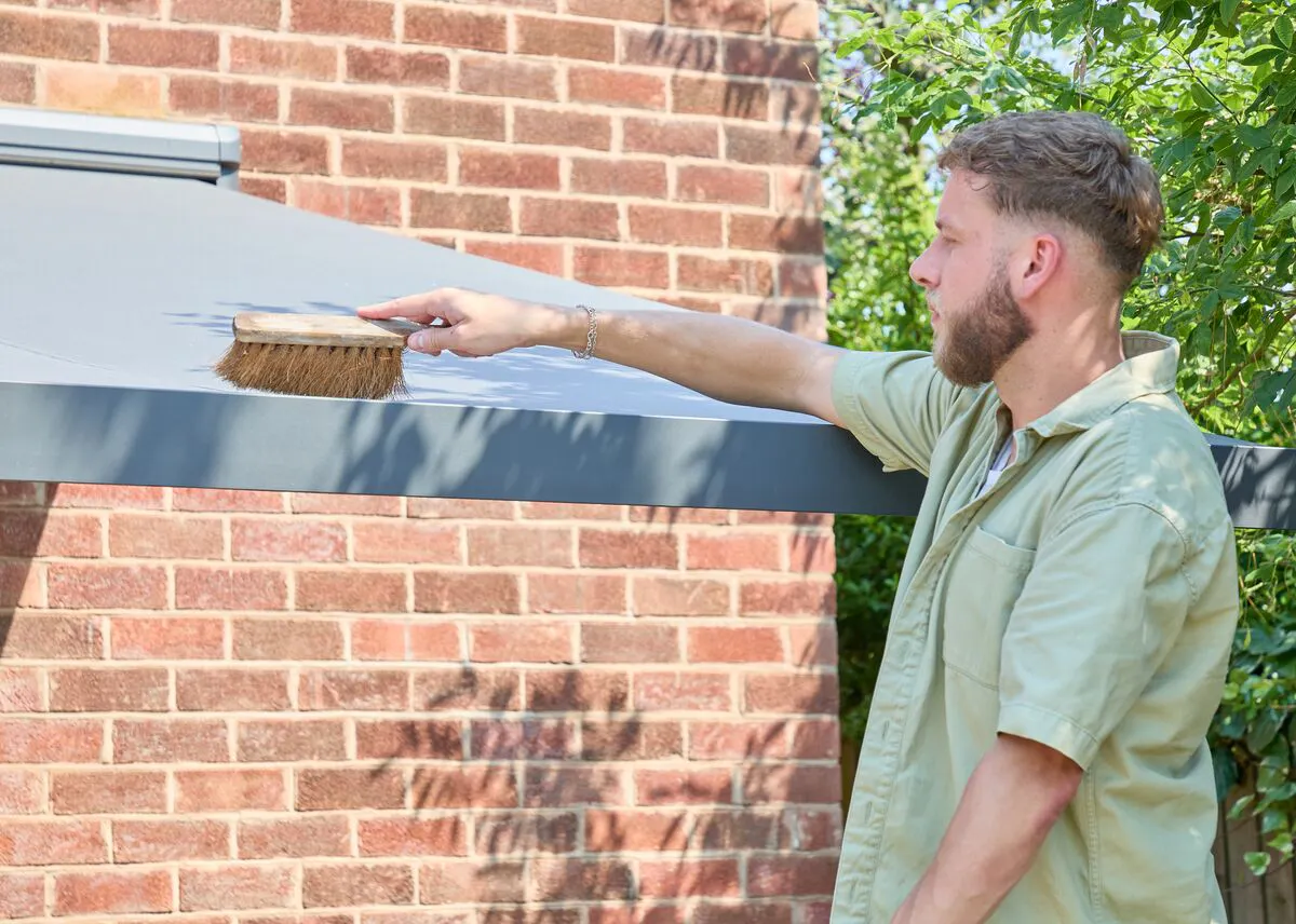 Person brushing debris from an extended grey patio awning attached to a brick house during outdoor cleaning and maintenance.
