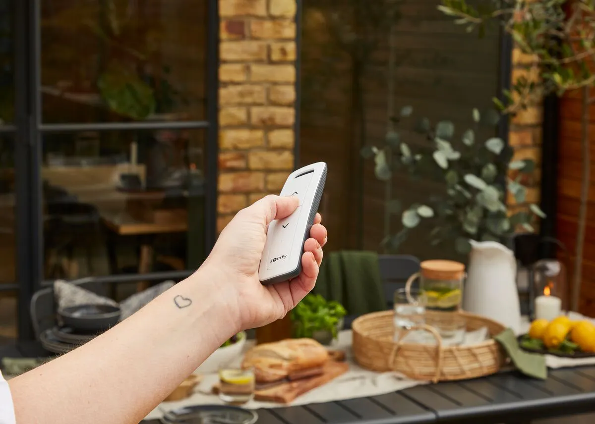 Hand using remote to operate a patio awning, with outdoor table setting and brick wall in the background.