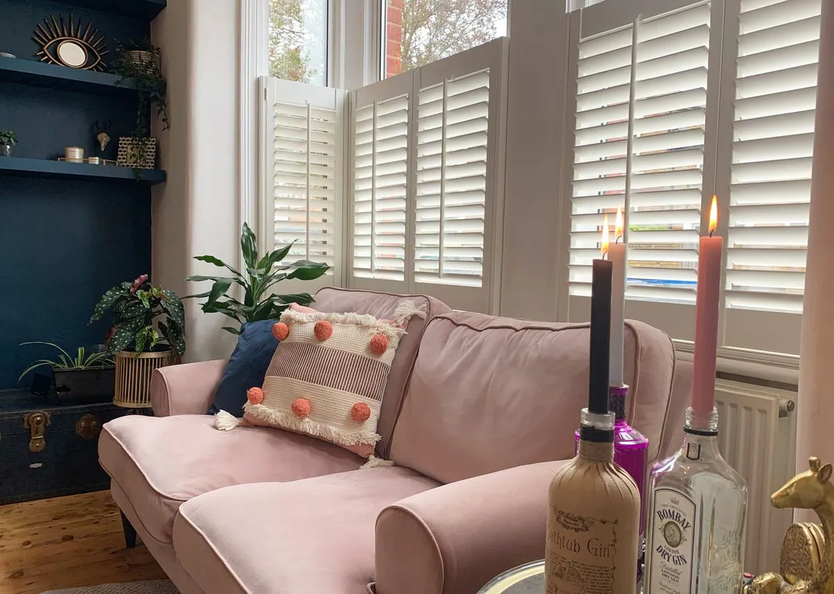 Soft living space with a blush sofa beneath a bright window fitted with white shutters, allowing gentle daylight to filter across plants and décor.