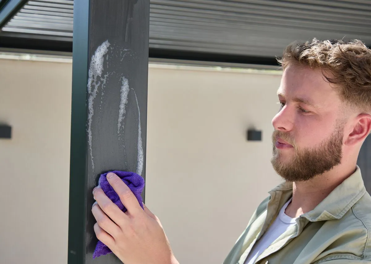Person wiping down the support post of a modern aluminium pergola with a cloth during outdoor cleaning and maintenance.