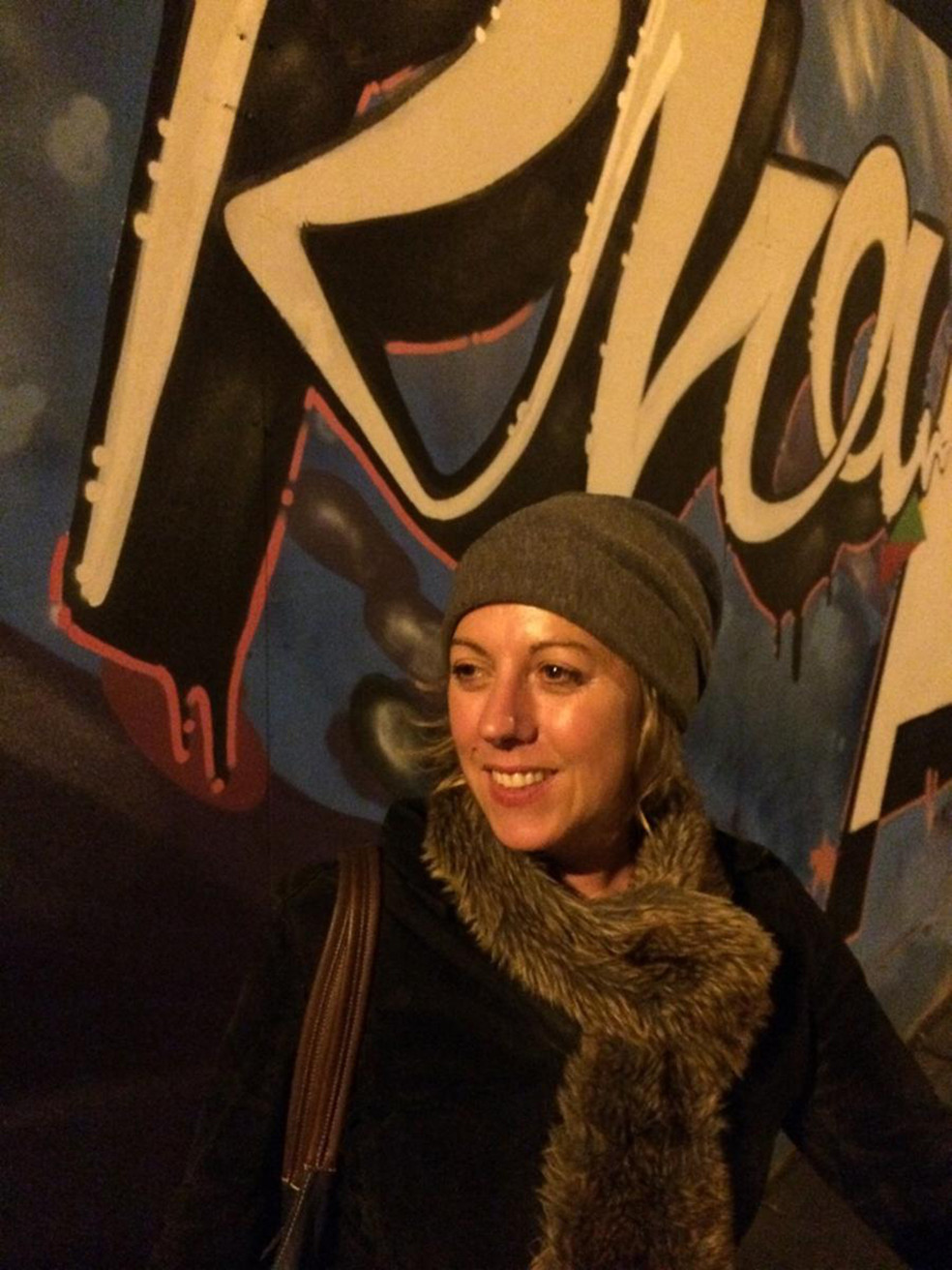 Person in a knit hat and thick scarf stands against a graffiti-covered wall with bold lettering, shoulder bag strap visible.