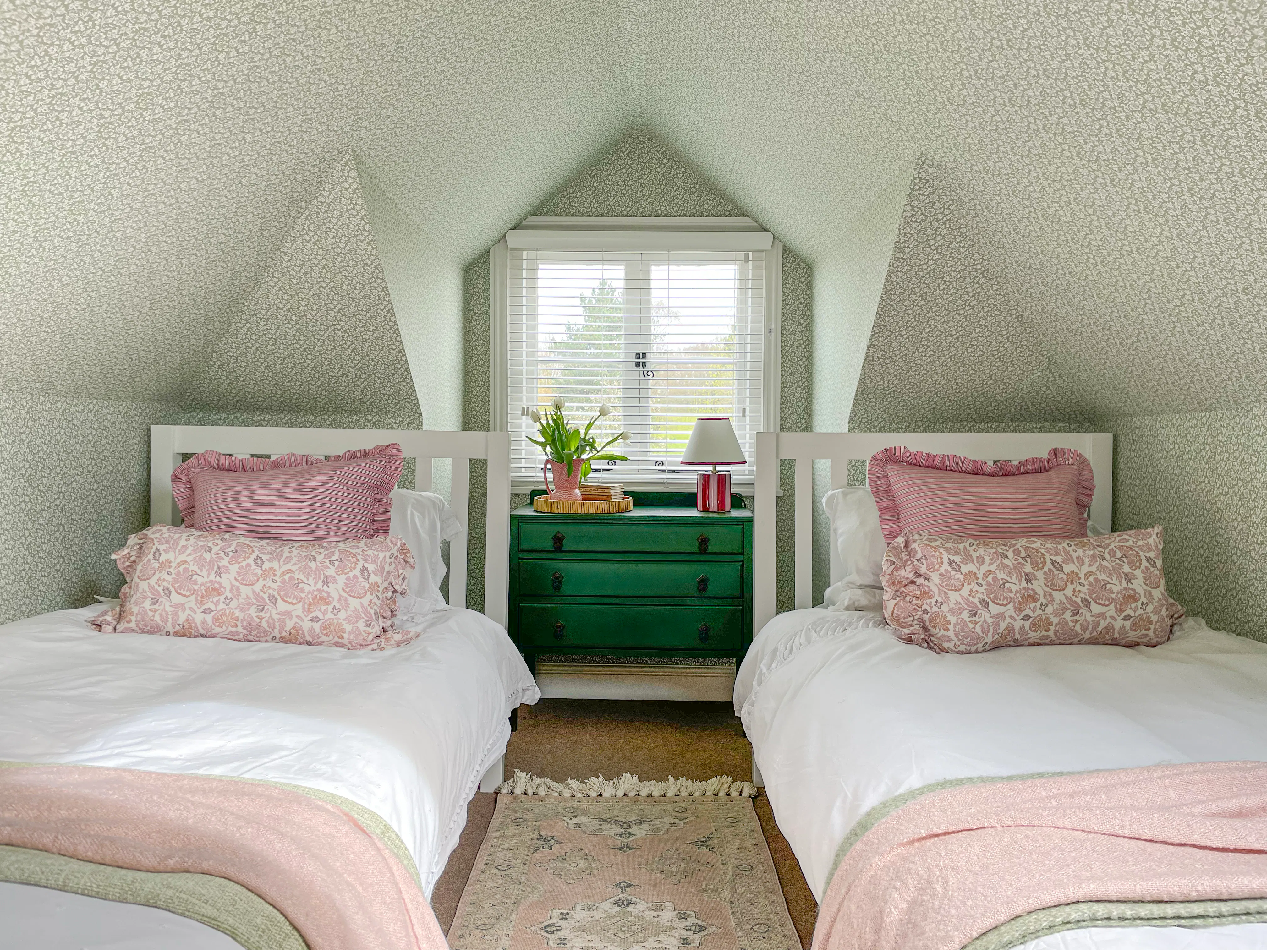 A charming twin bedroom with sage green botanical wallpaper covering the walls and sloped ceiling, white Venetian blind and pink and green coordinating bedding.