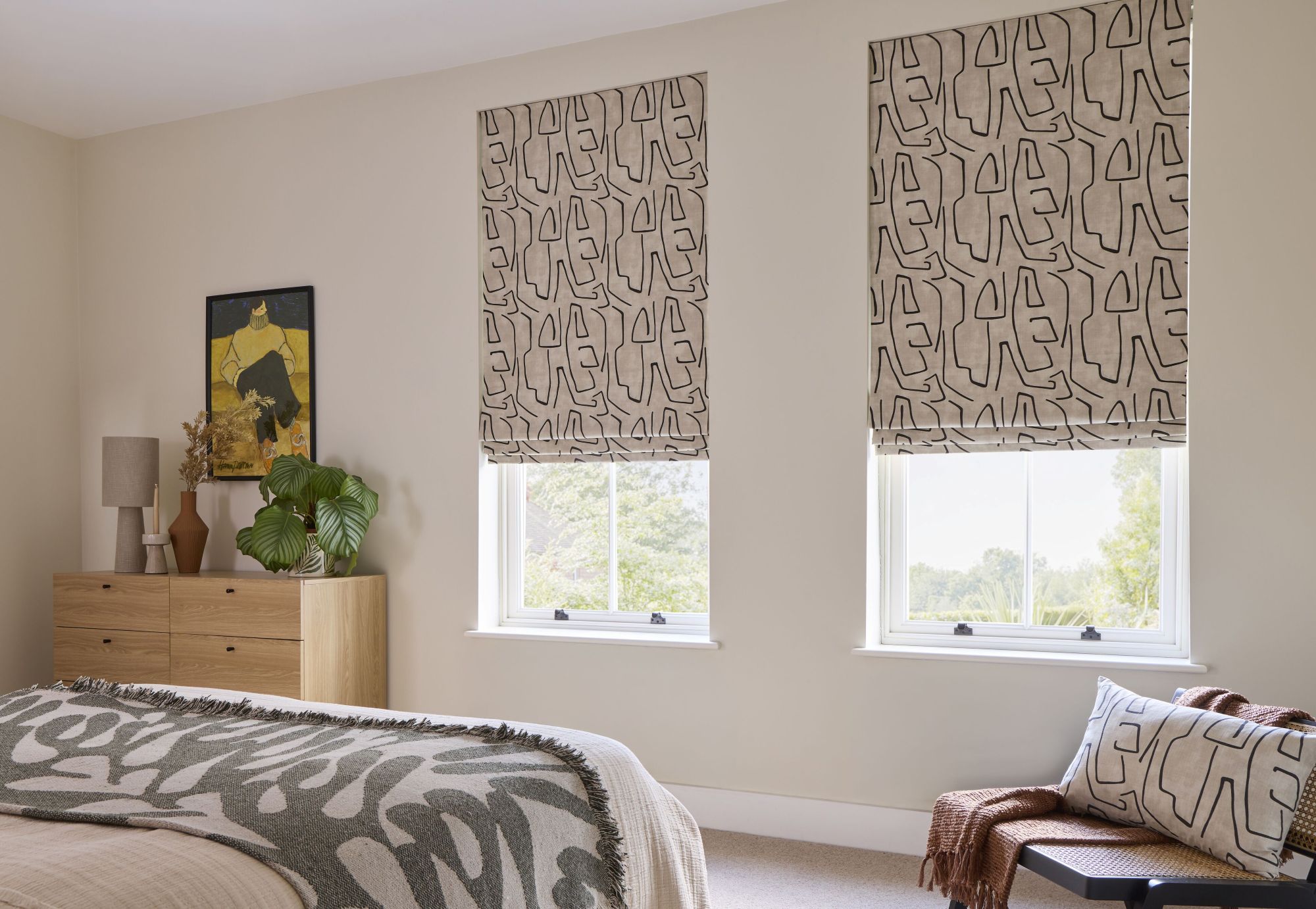 Geometric patterned Roman blinds in a neutral bedroom with soft furnishings, wooden furniture and large windows.