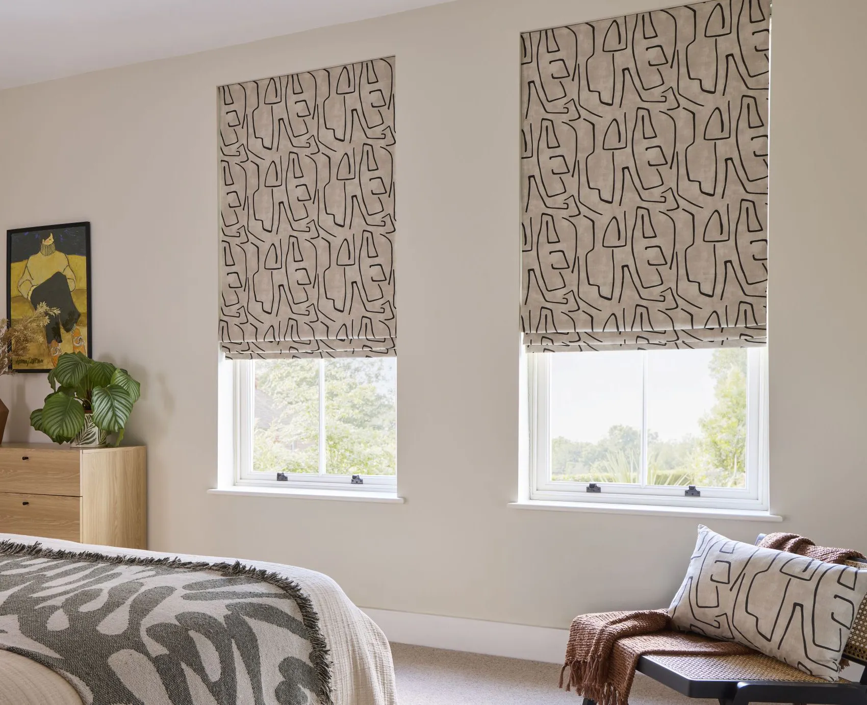 Geometric patterned Roman blinds in a neutral bedroom with soft furnishings, wooden furniture and large windows.