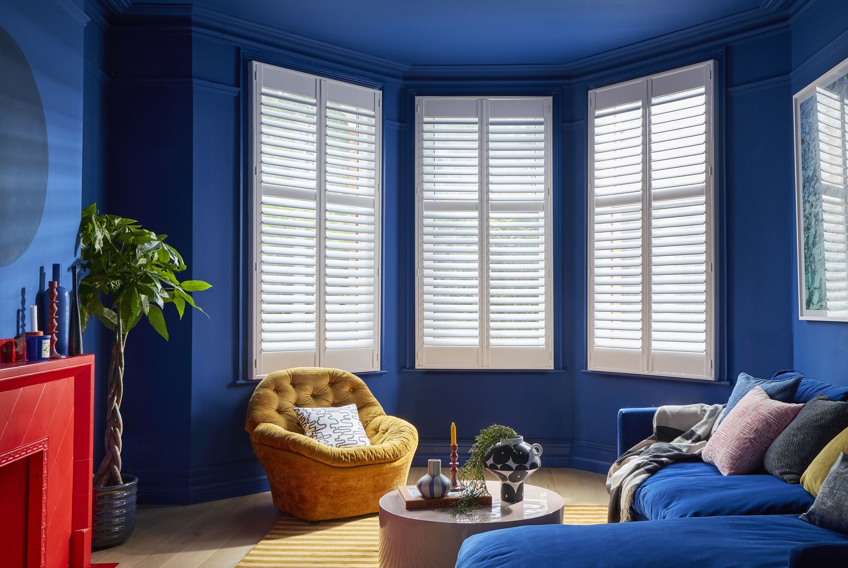 White full-height shutters fitted to a bay window in a bold blue living room with contemporary furniture and natural light.
