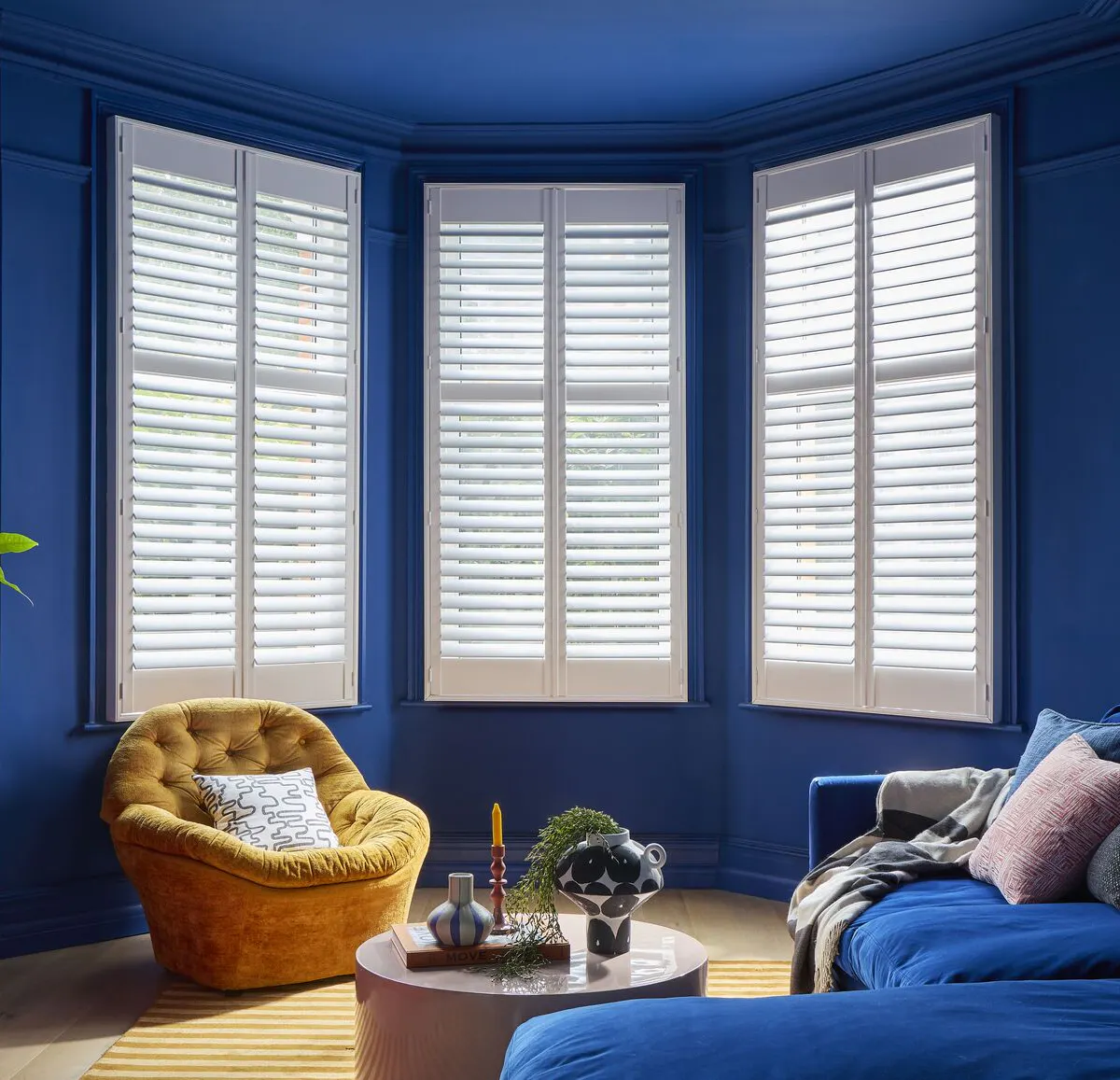 White full-height shutters fitted to a bay window in a bold blue living room with contemporary furniture and natural light.