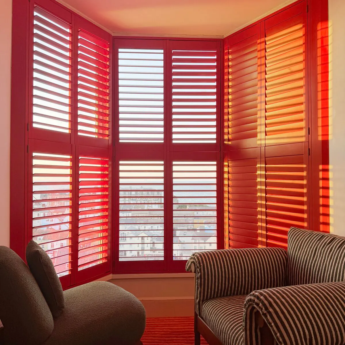 Red shutter panels across a bay window, filtering warm sunlight into a cosy seating area with striped armchairs and a textured rug.