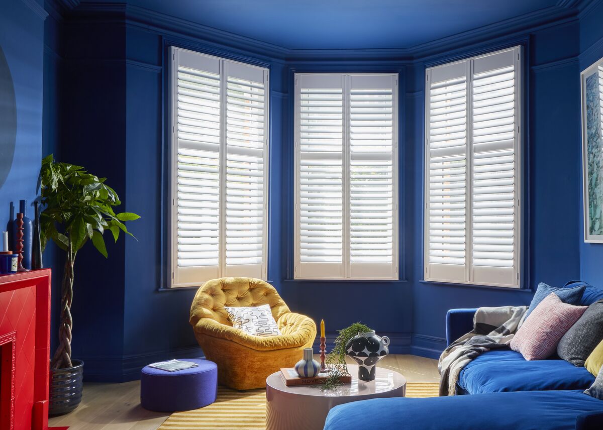 A colour-drenched blue living room with a large blue velvet corner sofa facing a red-painted fireplace. A bay window is framed with white plantation shutters.