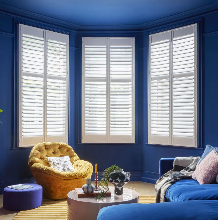 A colour-drenched blue living room with a large blue velvet corner sofa facing a red-painted fireplace. A bay window is framed with white plantation shutters.