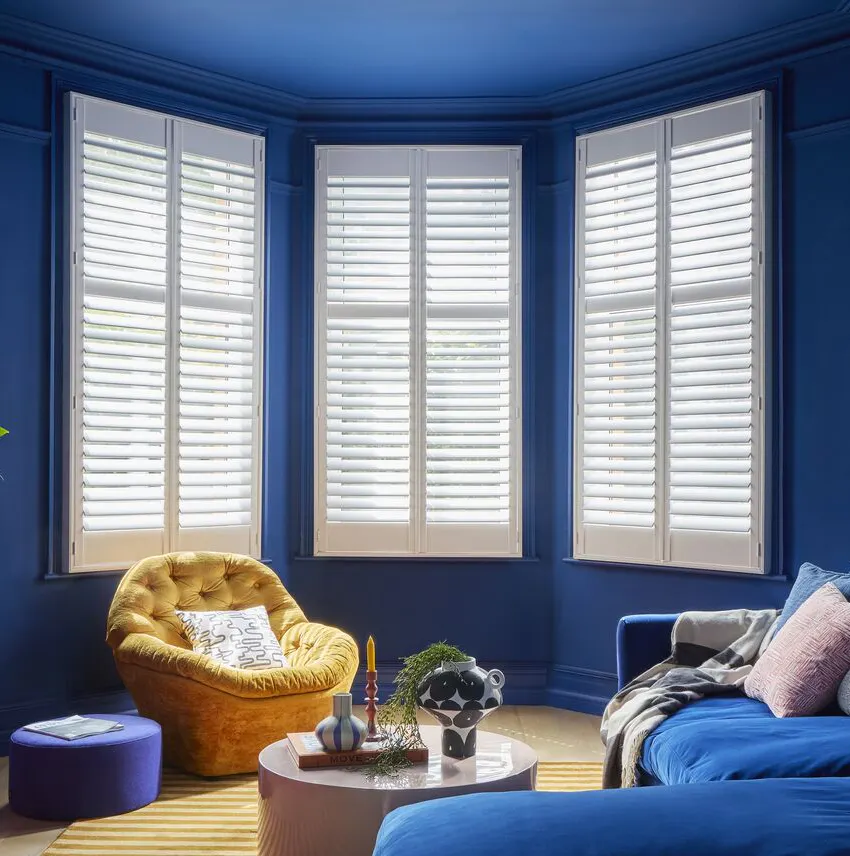 A colour-drenched blue living room with a large blue velvet corner sofa facing a red-painted fireplace. A bay window is framed with white plantation shutters.