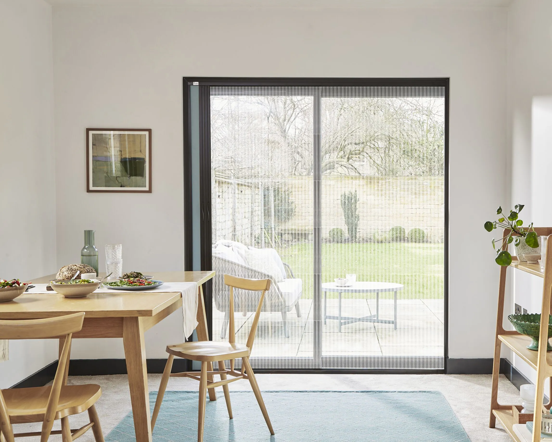Dining area with large sliding glass doors fitted with a discreet Fly Screen, opening onto a green garden and bringing daylight into the calm space. 