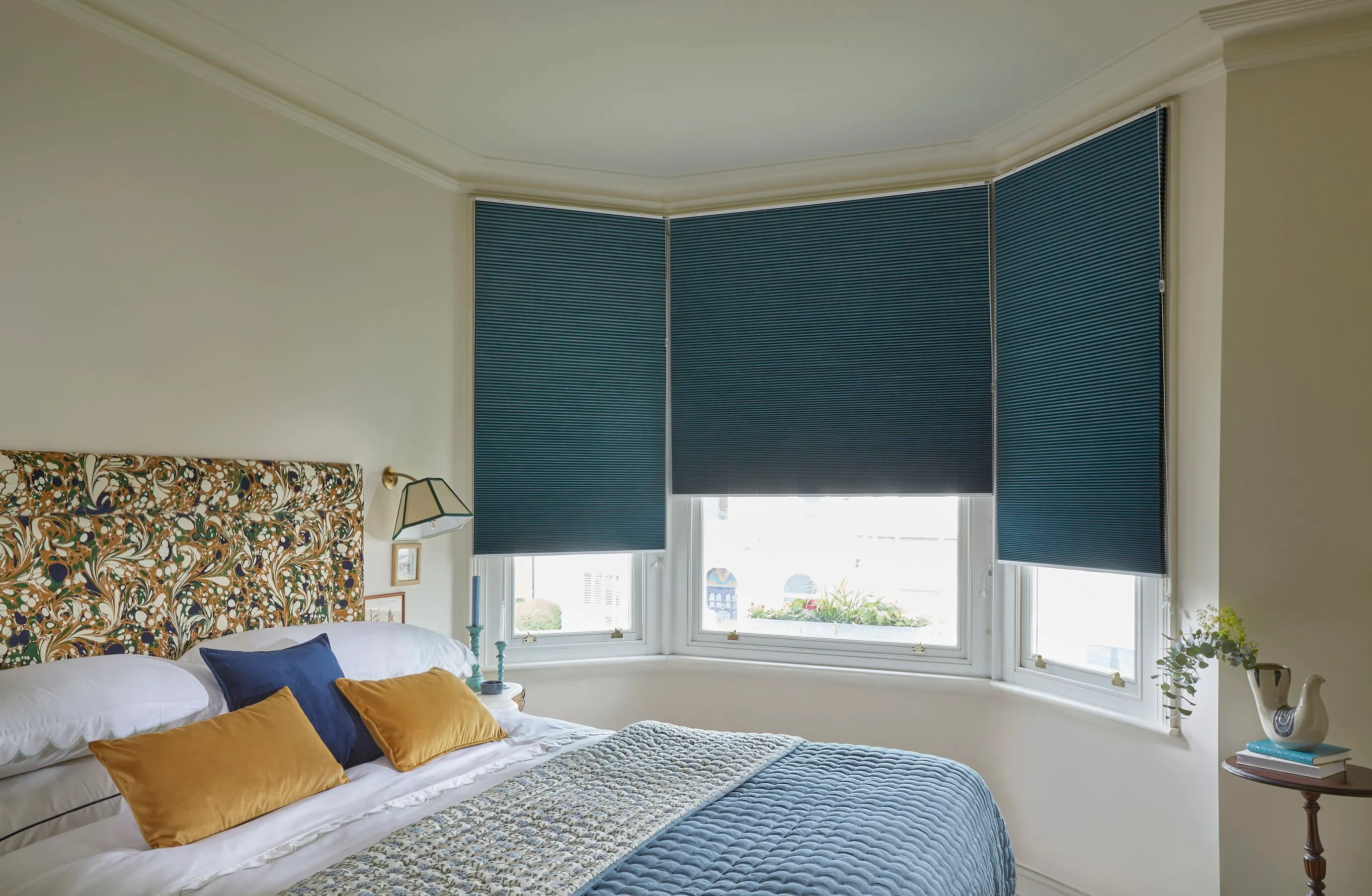 Dark blue Duette blinds cover a bay window, adding privacy and light control to a bright bedroom with patterned headboard and blue bedding.