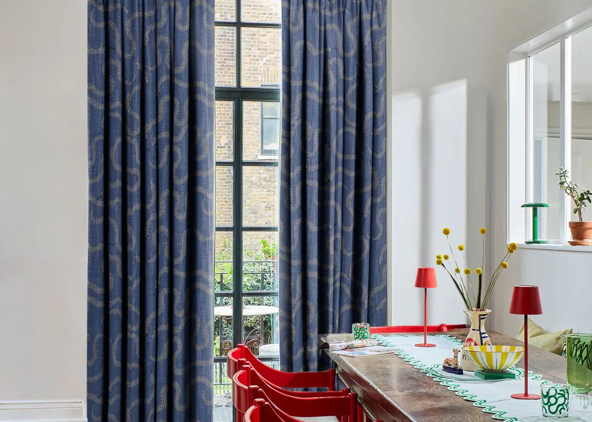 Tall black-framed window dressed with blue patterned curtains, bringing light into a dining space with colourful furnishings.