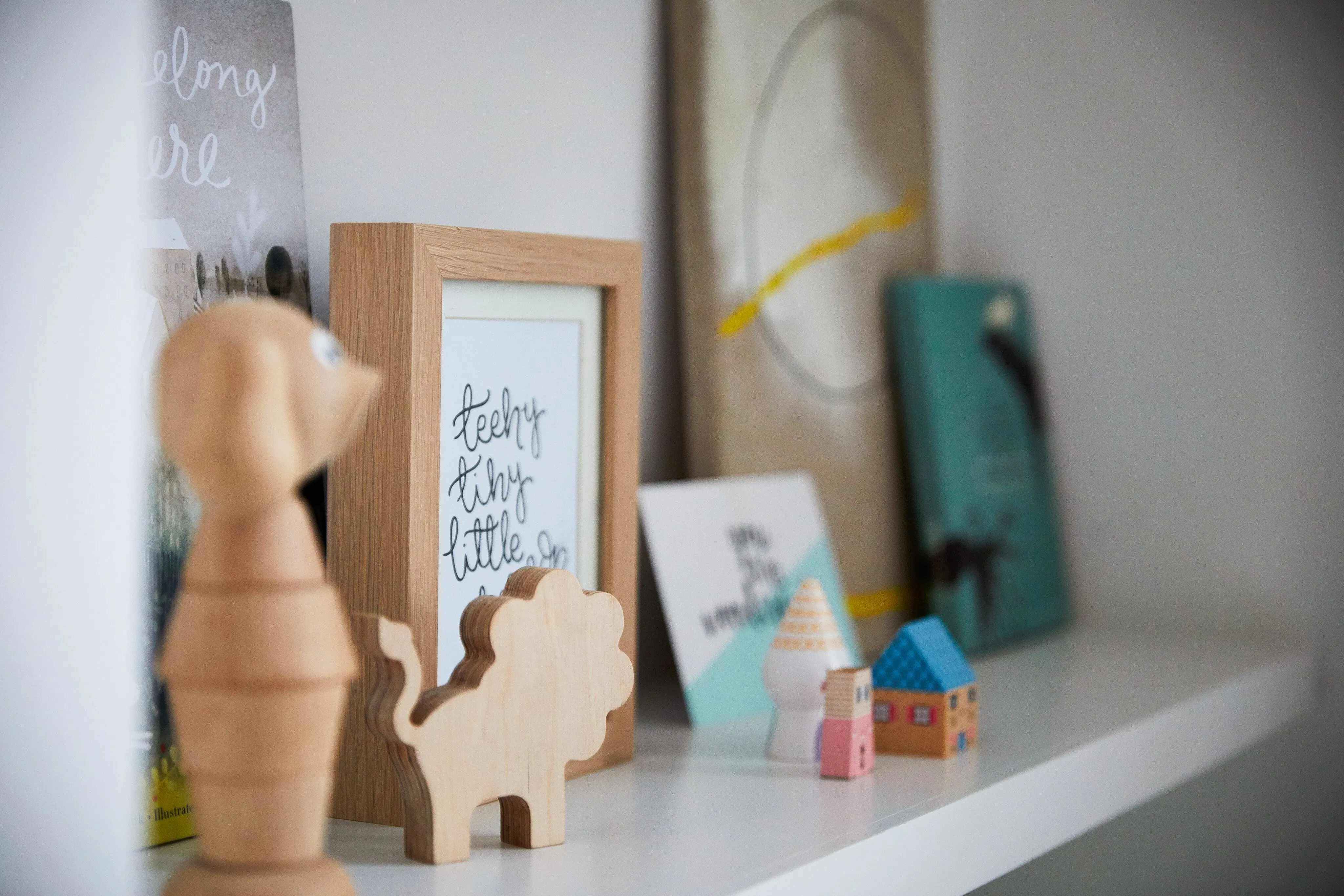 White shelf styled with framed prints, wooden ornaments and miniature houses, shown in soft focus against a neutral wall.