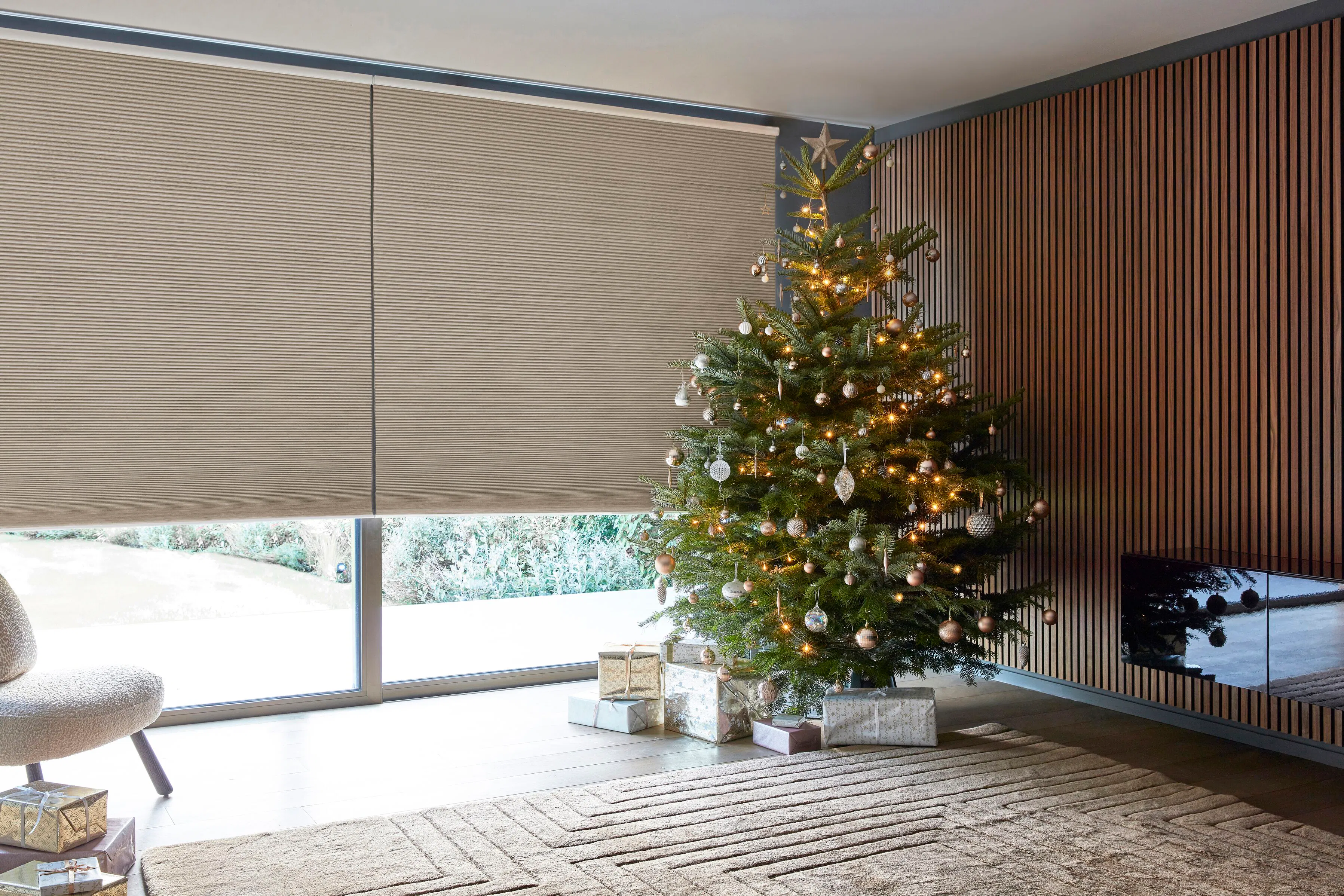 A modern living room with taupe honeycomb blinds and a wood-slatted feature wall. A natural Christmas tree decorated in copper and gold baubles stands on a textured neutral rug.