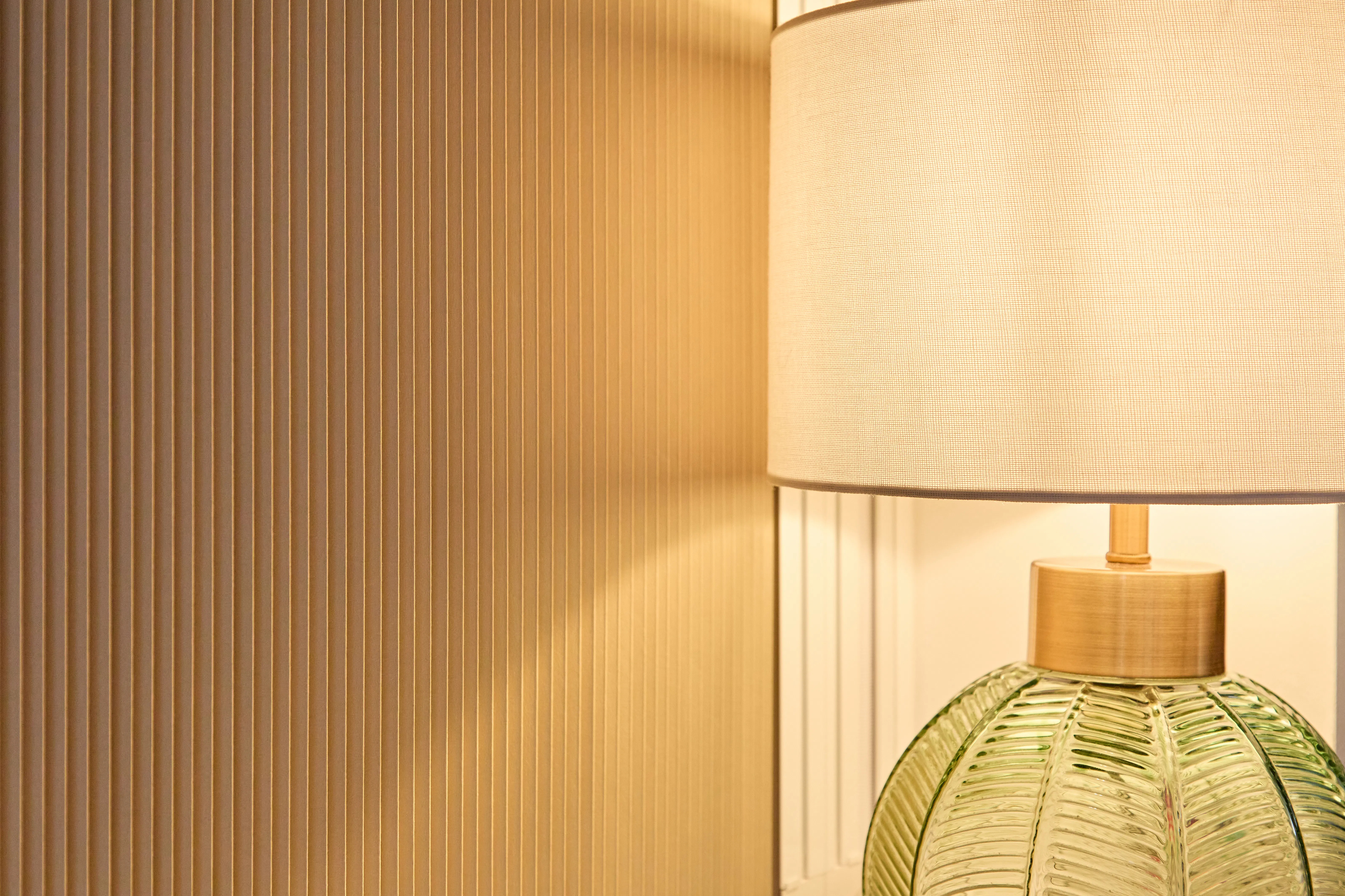 A close-up of a warm-toned pleated blind beside a table lamp with a ribbed green glass base and linen shade.