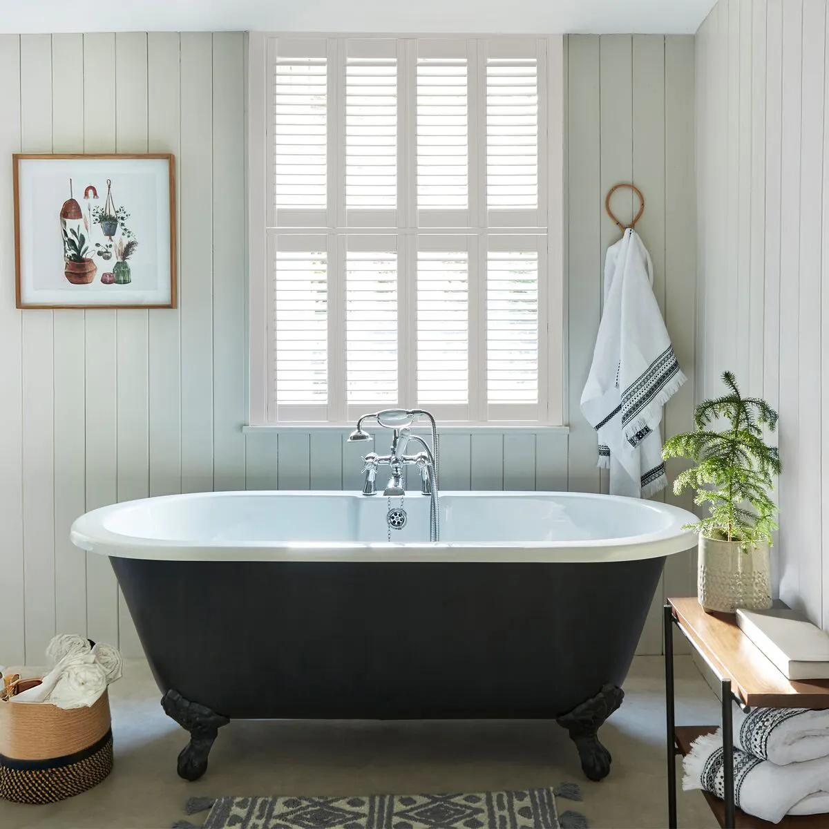 Pale green tongue-and-groove panelled bathroom with a black roll-top bath, white shutters and woven baskets.