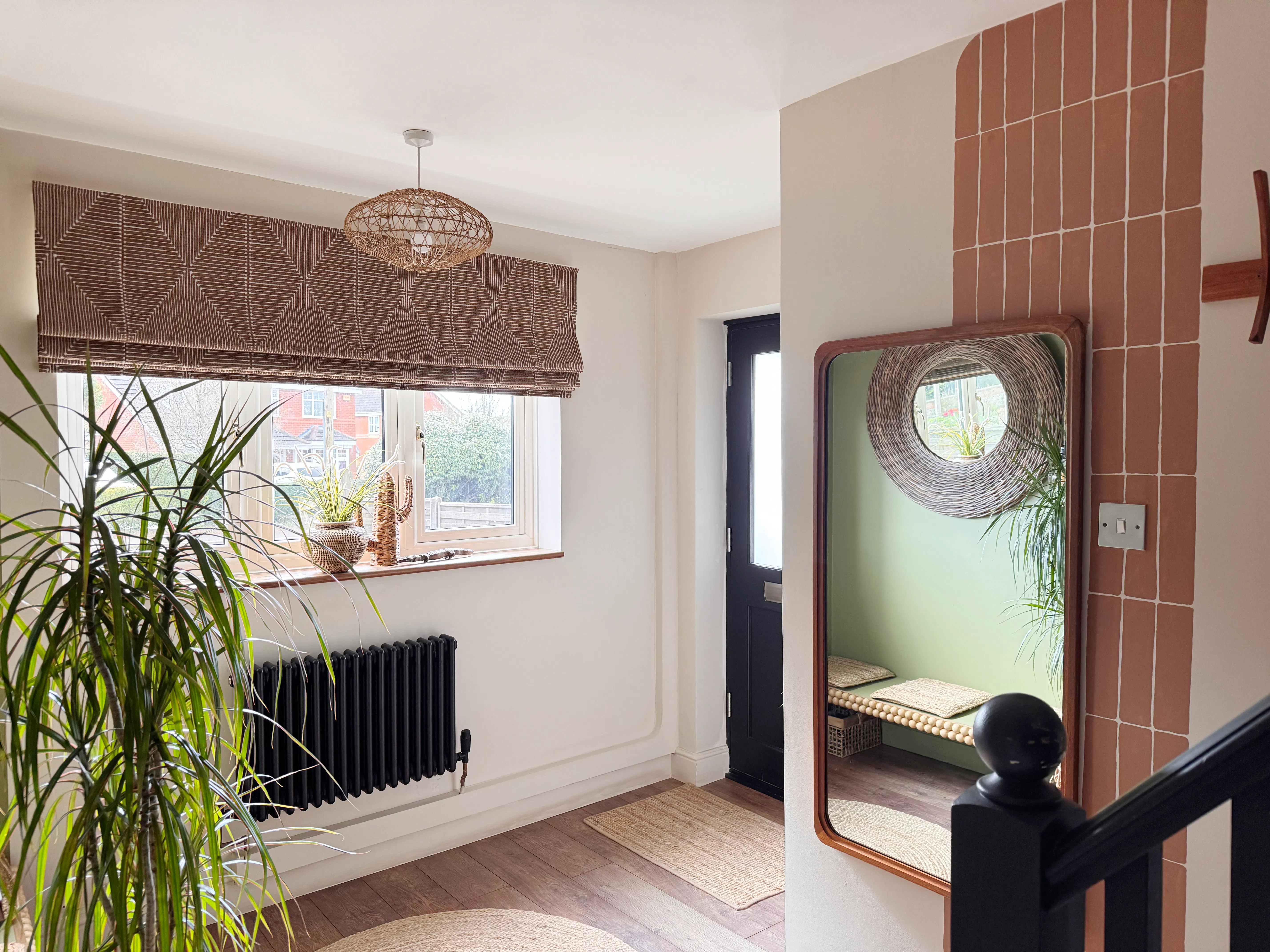 Bright hallway with front window dressed in a Roman blind, black radiator below, indoor plants, wall mirror and warm tiled feature wall.