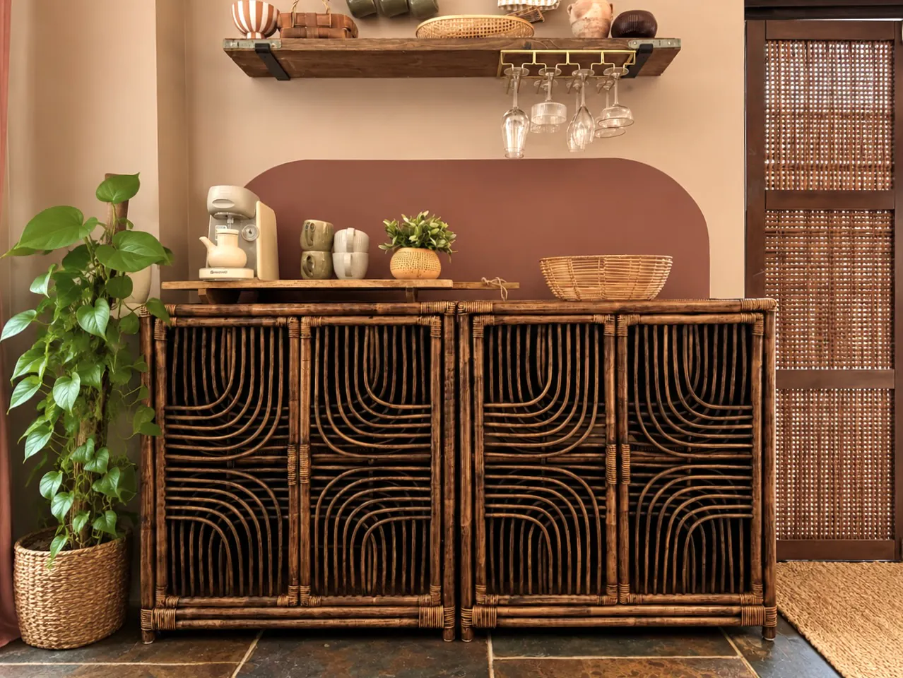 Boho-style interior with rattan sideboard, woven sliding door, coffee station on open shelves, hanging glassware and leafy plant accents.