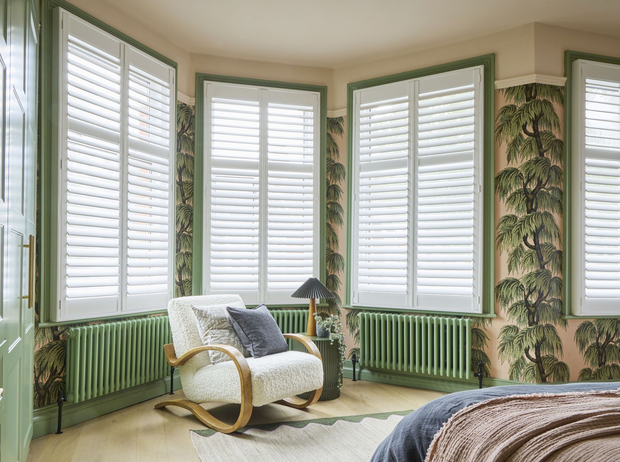 White full-height shutters installed across a bay window in a stylish bedroom with botanical wallpaper and green accents.