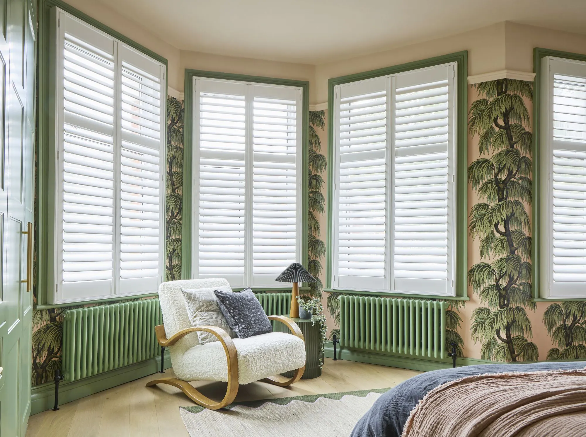 White full-height shutters installed across a bay window in a stylish bedroom with botanical wallpaper and green accents.