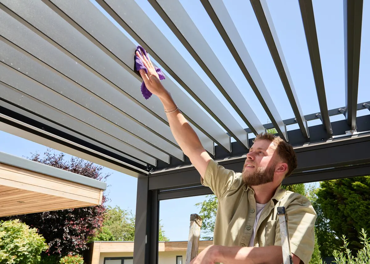 Person cleaning the louvred roof of a modern aluminium pergola with a cloth, reaching up from a stepladder on a sunny day.