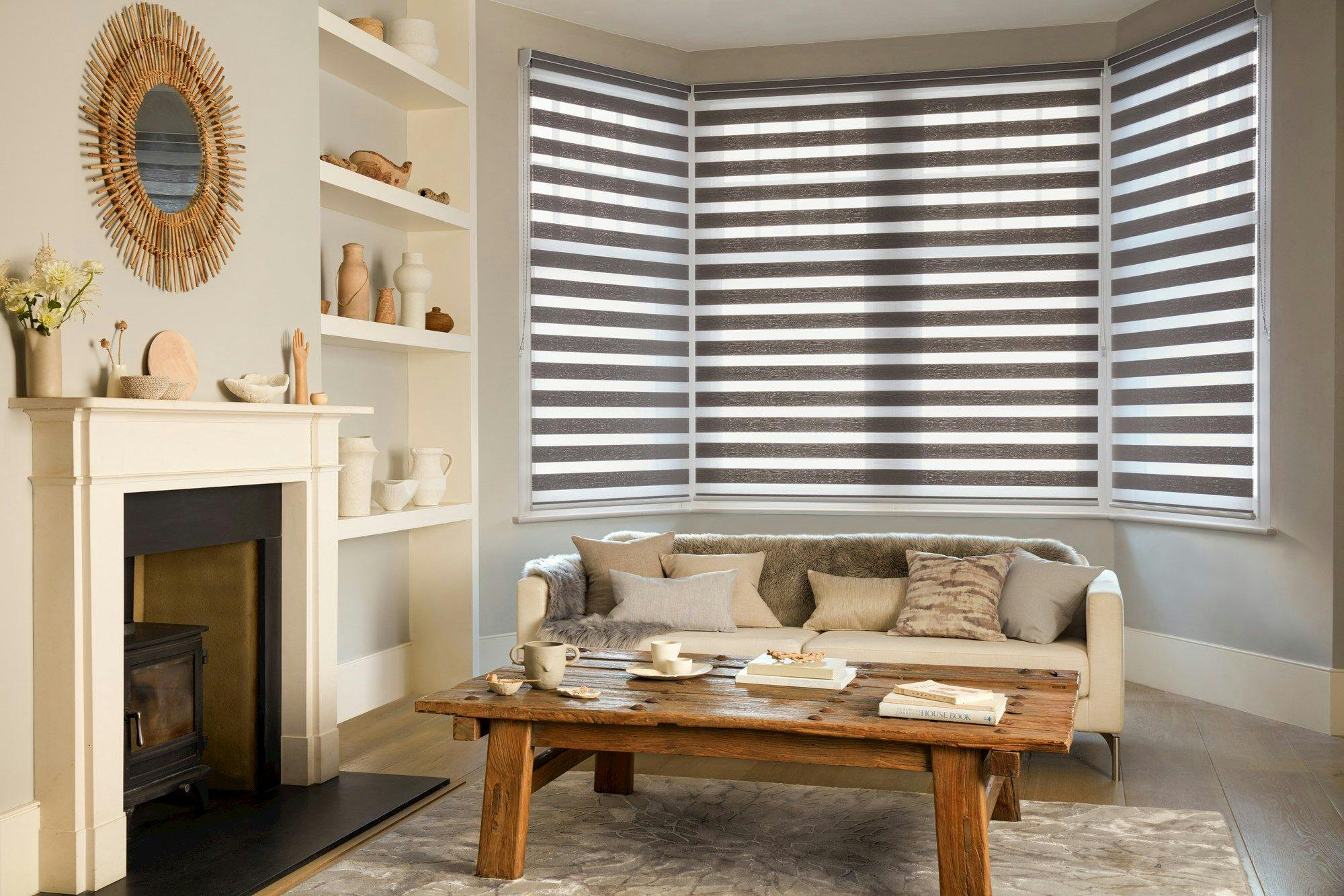 A cozy living room features a beige sofa with cushions, a wooden coffee table, a decorative fireplace, and striped blinds in a bay window, set with neutral tones and minimalist decor.
