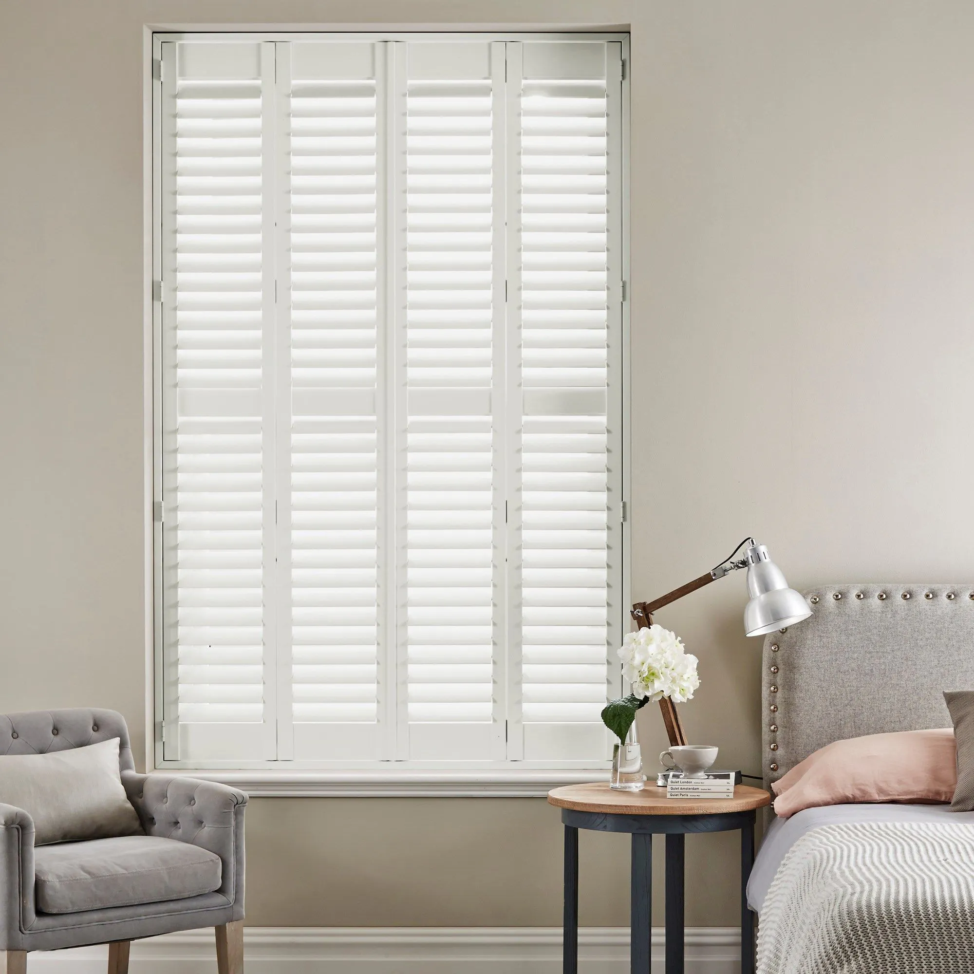 bright white shutters next to grey bed with pink pillow and flower and lamp on bedside table