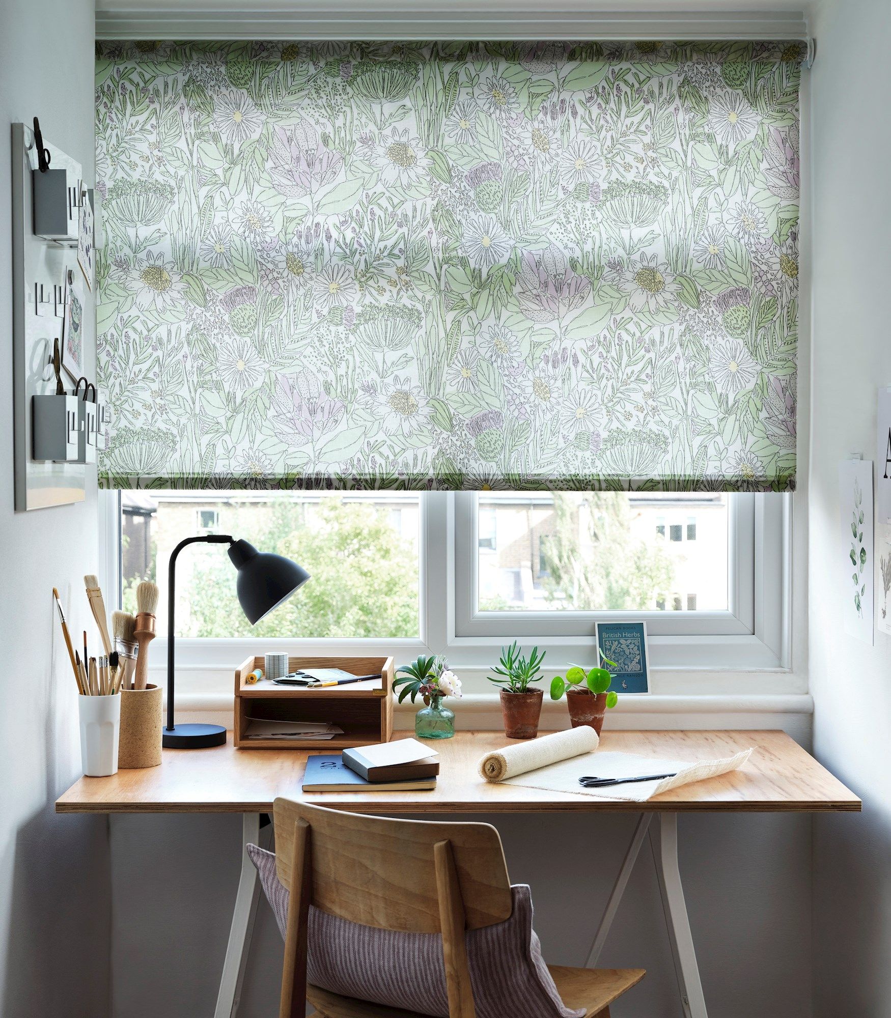 A wooden desk holds a lamp, notebooks, art supplies, and small potted plants. Floral-patterned blinds cover the window, and wall-mounted organizers hang nearby, providing a tidy workspace.