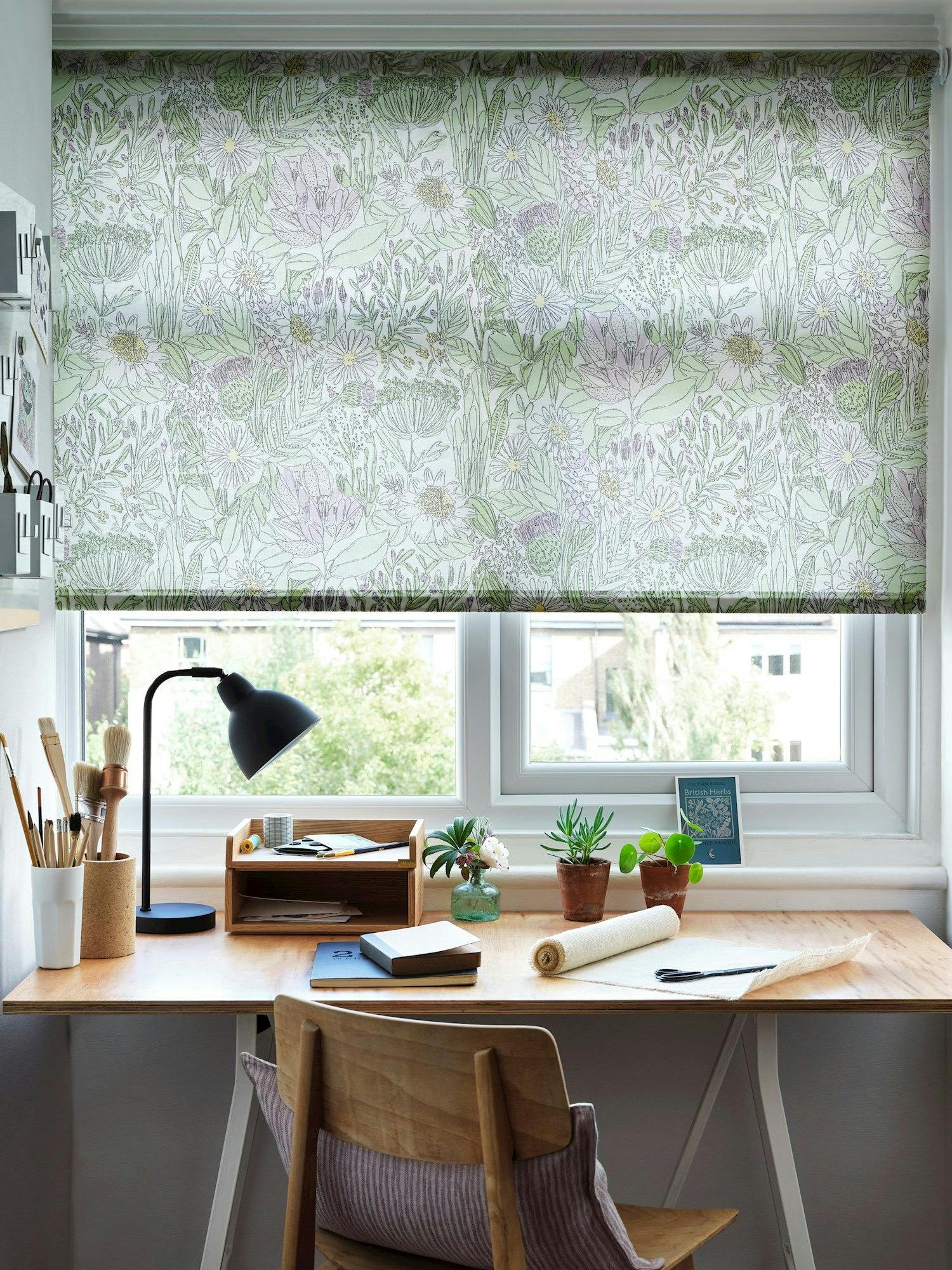 A wooden desk holds a lamp, notebooks, art supplies, and small potted plants. Floral-patterned blinds cover the window, and wall-mounted organizers hang nearby, providing a tidy workspace.