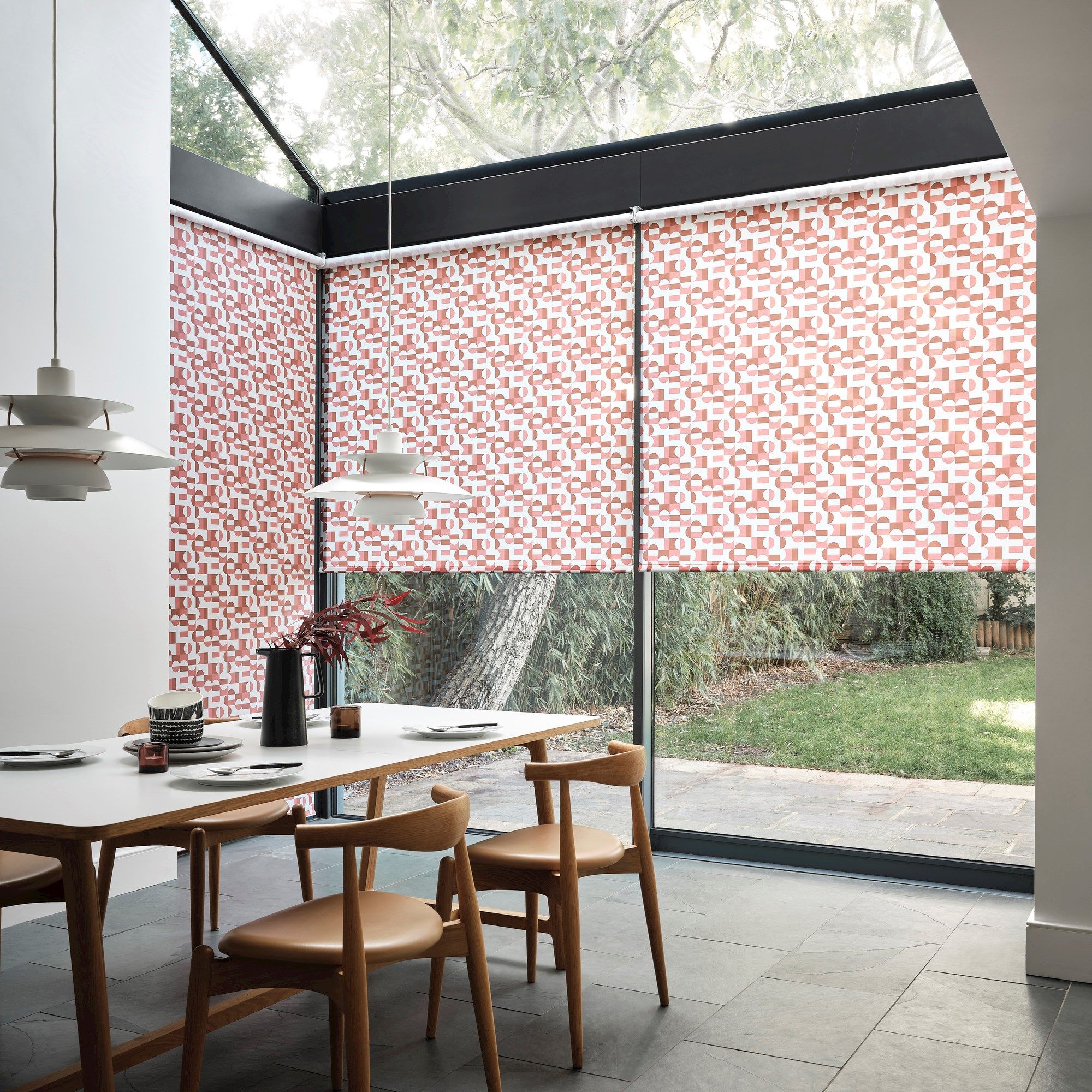 A dining table with wooden chairs holds tableware and a vase. Geometric roller blinds cover large windows, with lush greenery visible outside. Modern pendant lights hang above.