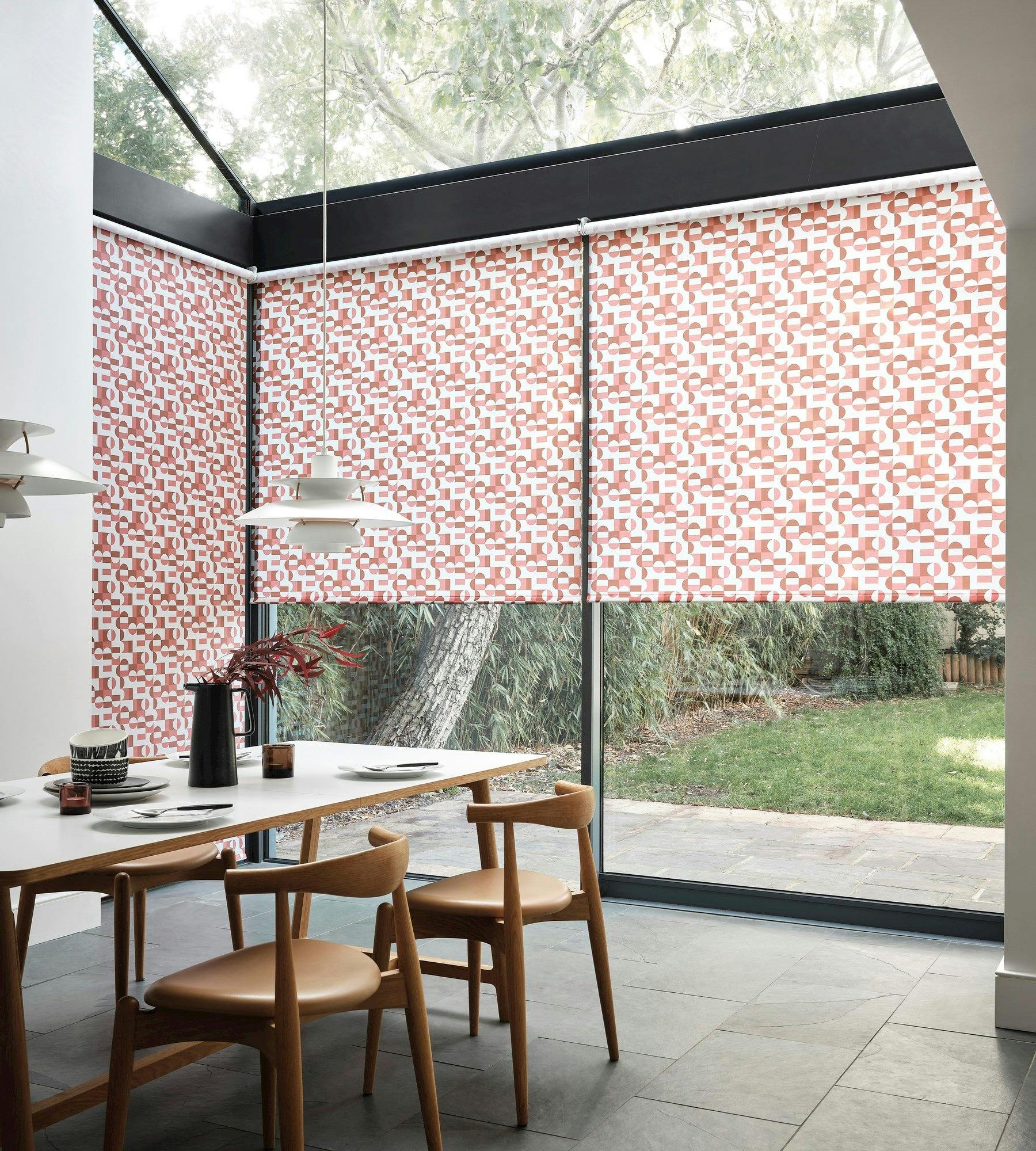 A dining table with wooden chairs holds tableware and a vase. Geometric roller blinds cover large windows, with lush greenery visible outside. Modern pendant lights hang above.