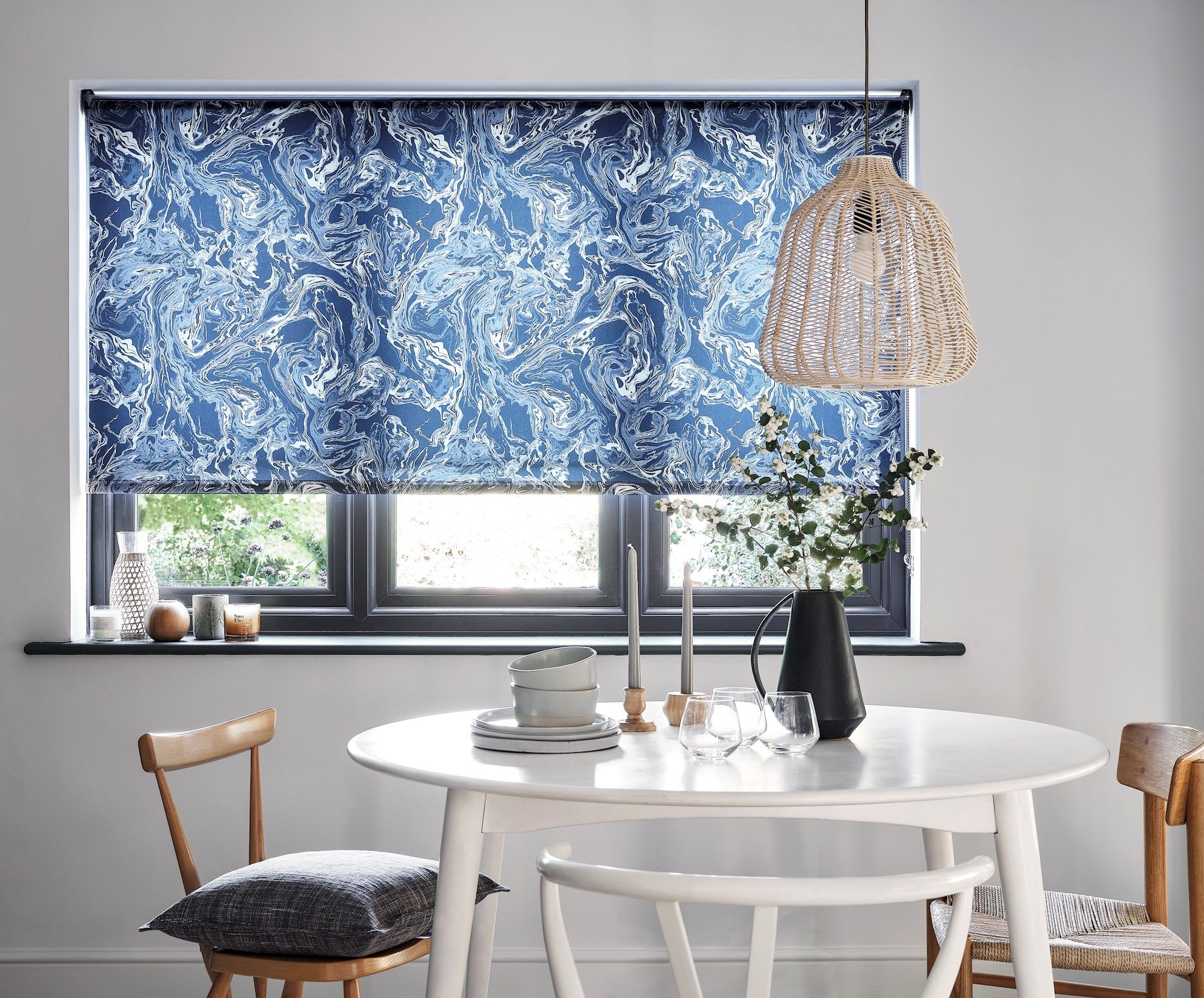 A blue, marbled-patterned window blind partially unrolls in a cozy dining area. It hangs above a round white table set with dishes and a vase, beneath a woven rattan pendant light.