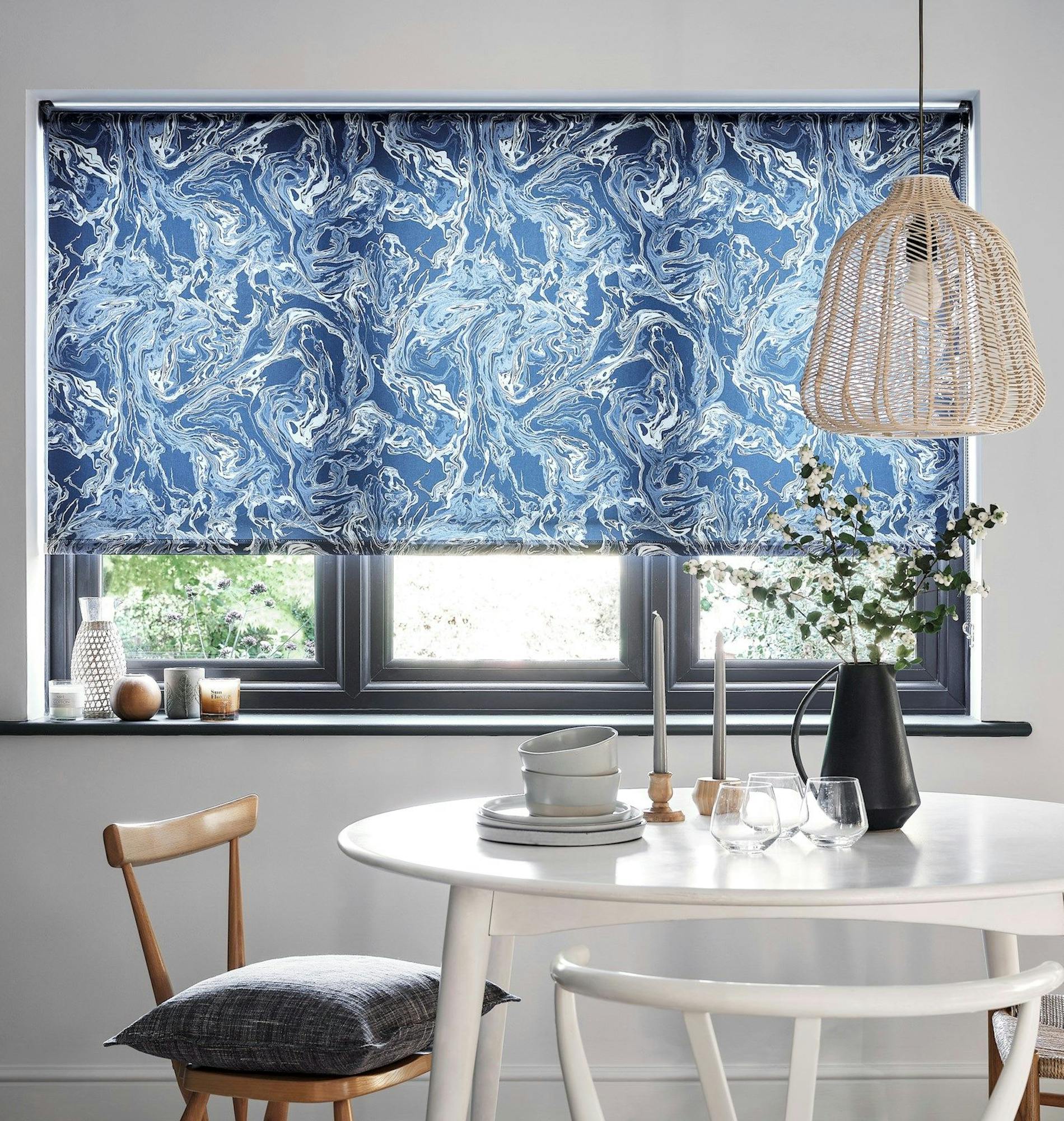 A blue, marbled-patterned window blind partially unrolls in a cozy dining area. It hangs above a round white table set with dishes and a vase, beneath a woven rattan pendant light.
