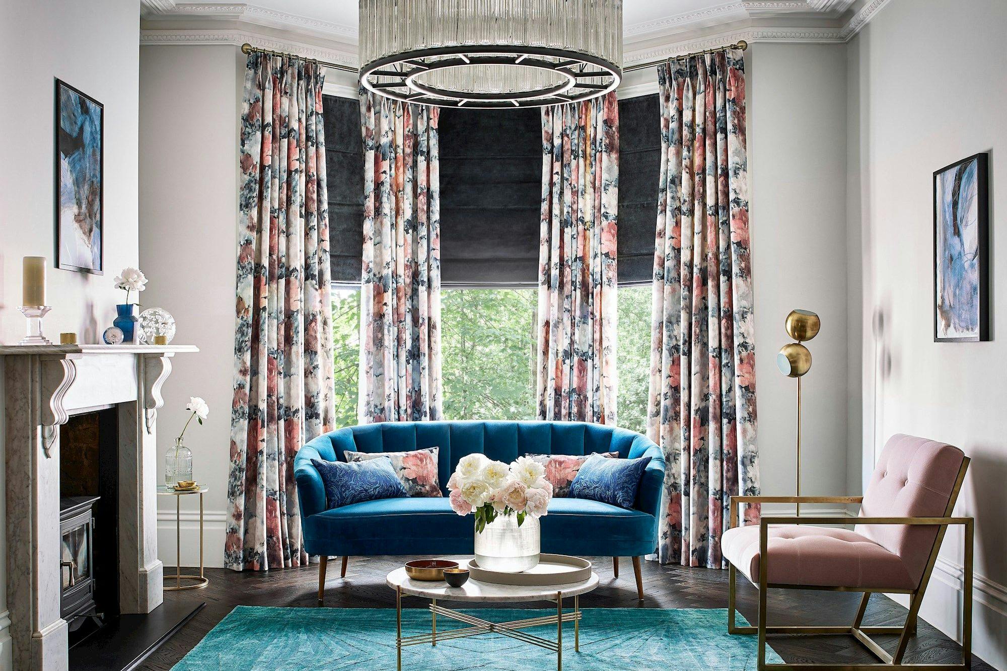 Dark grey velvet roman blind under floral print curtains in a bright living room with blue and rose pink accents