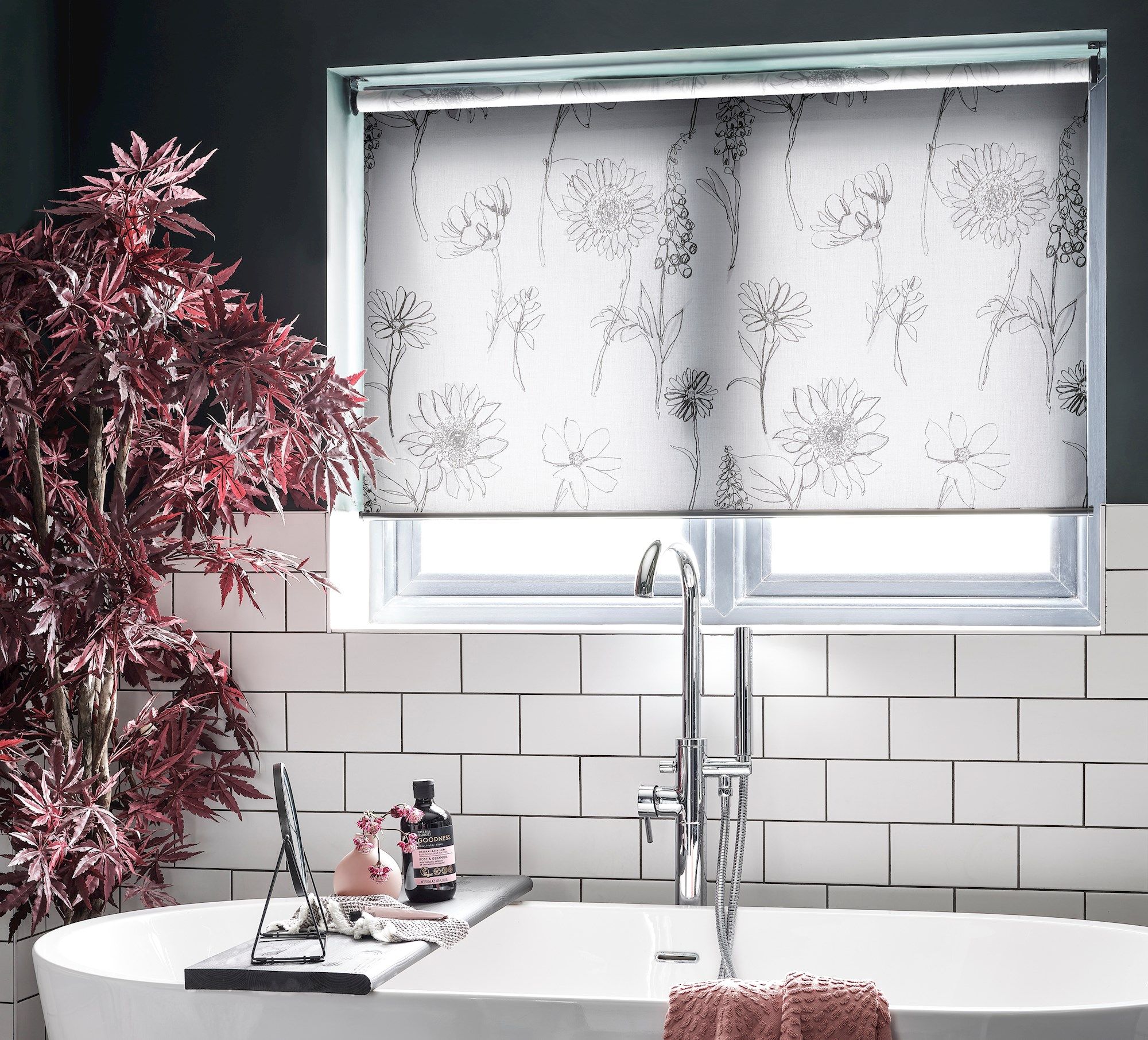 A bathtub with a faucet is set beneath a window, covered with floral-patterned blinds, surrounded by white tiles and a red plant, conveying a modern bathroom ambiance.