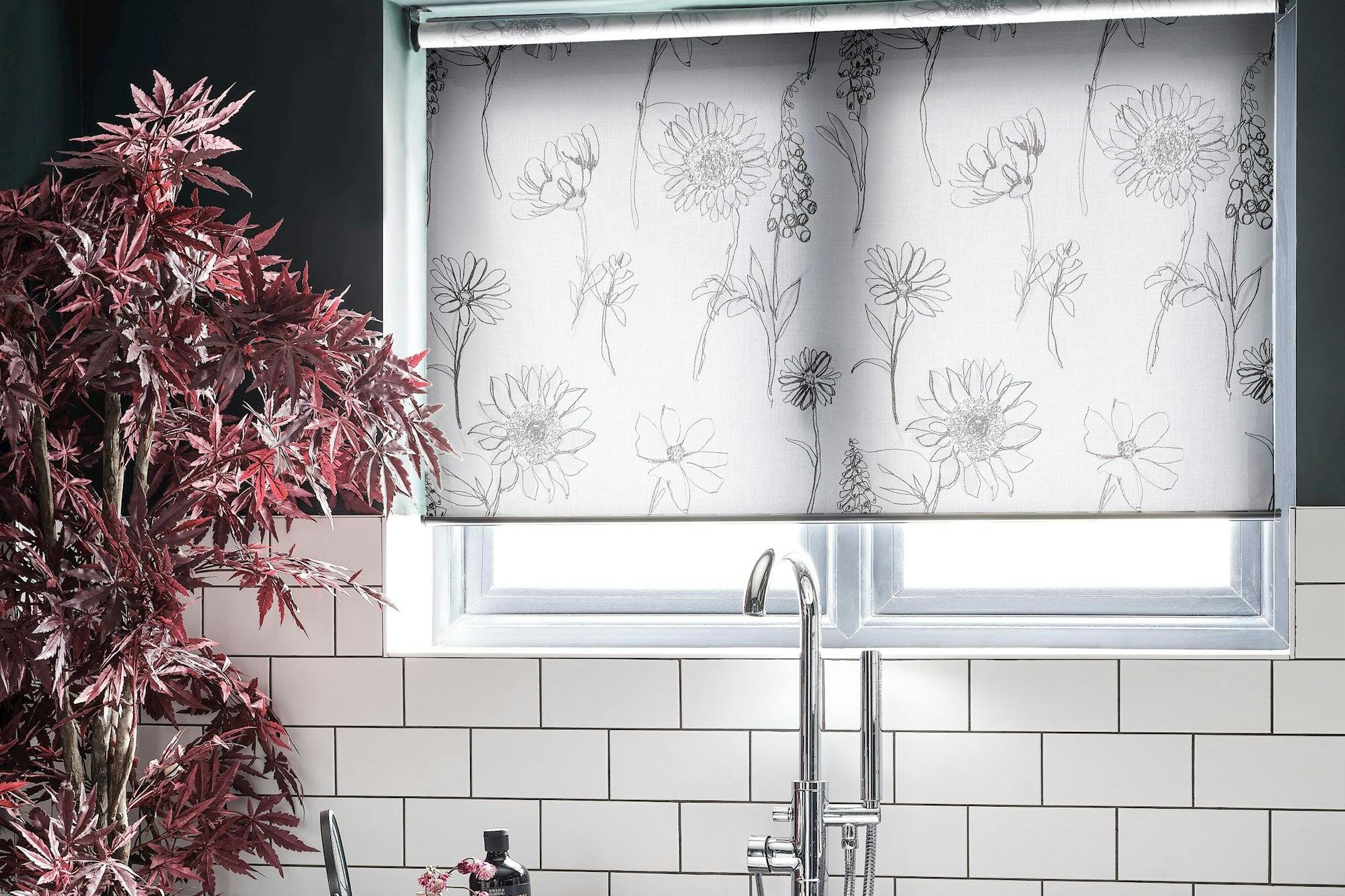 A bathtub with a faucet is set beneath a window, covered with floral-patterned blinds, surrounded by white tiles and a red plant, conveying a modern bathroom ambiance.