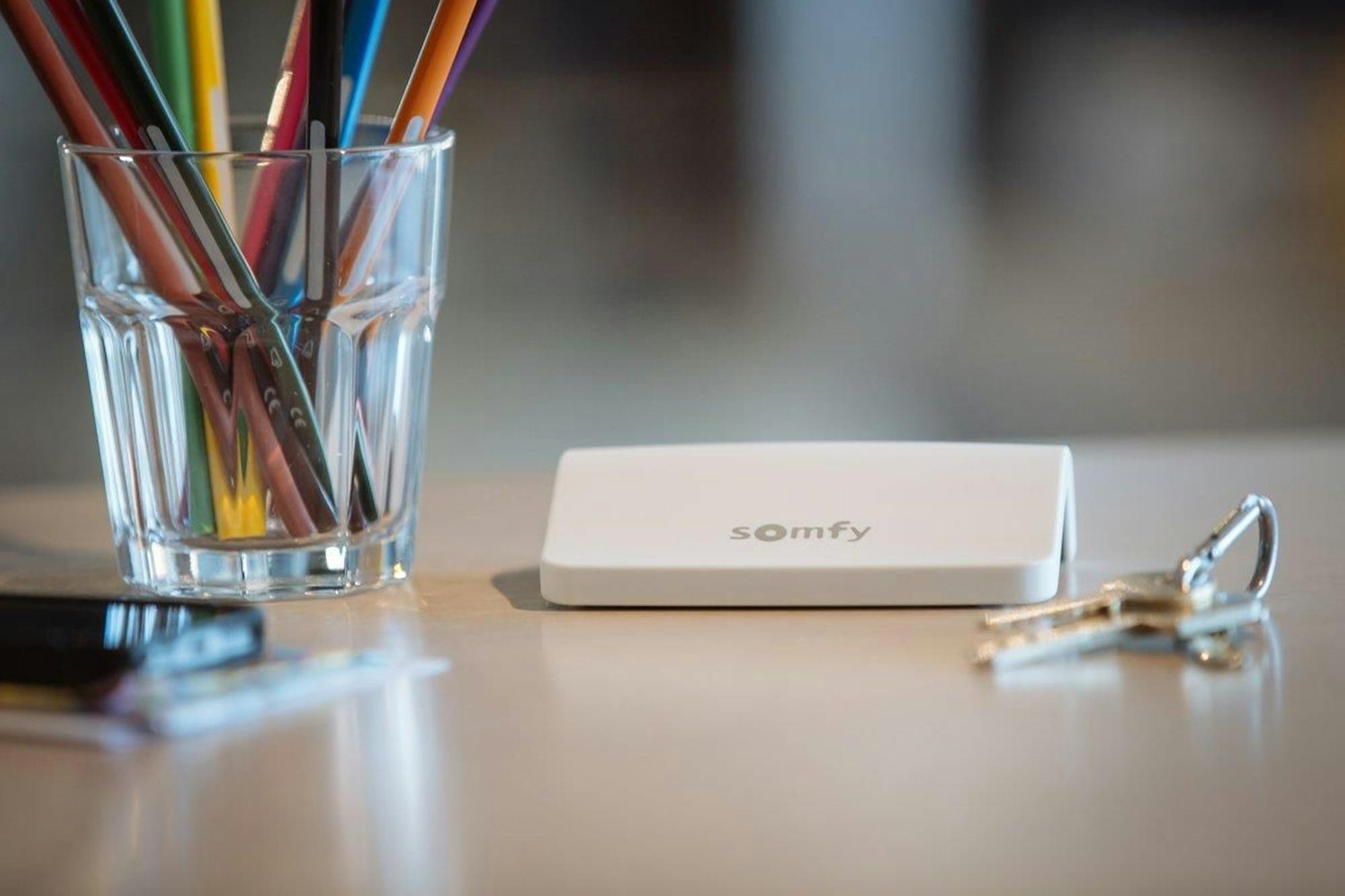 A white rectangular device labeled "somfy" rests on a wooden surface, accompanied by a clear glass holding colorful pencils and a set of metallic keys.
