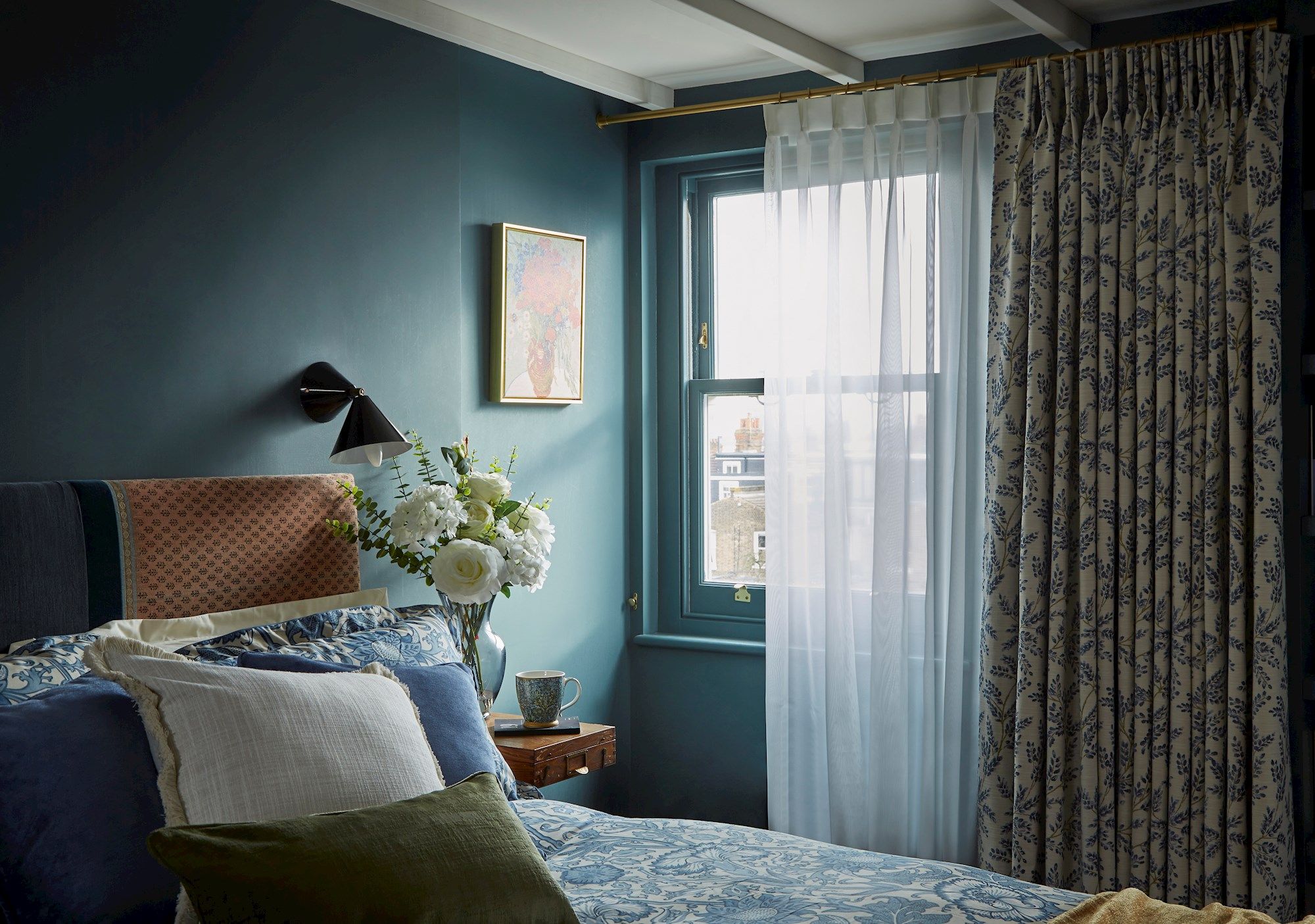 A bed adorned with patterned blue and white linens rests near a wooden side table with a vase of white flowers, under a window framed by sheer and floral curtains in a cozy room.