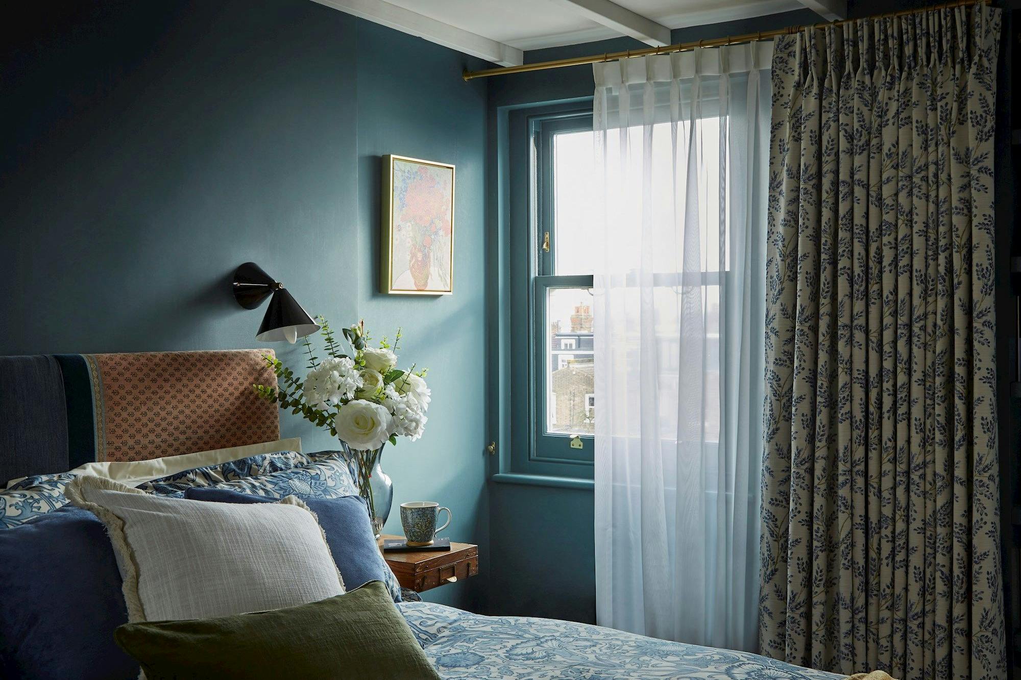 A bed adorned with patterned blue and white linens rests near a wooden side table with a vase of white flowers, under a window framed by sheer and floral curtains in a cozy room.