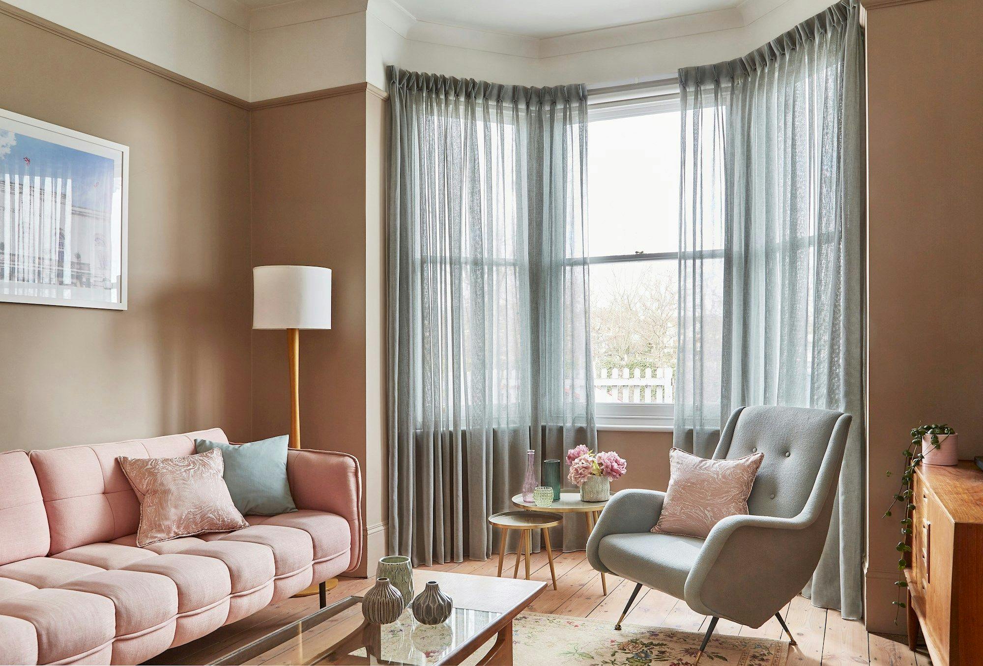 A pink sofa and an armchair with cushions sit in a cozy living room. Soft curtains frame a large window, with sunlight illuminating a coffee table and decorative vases.