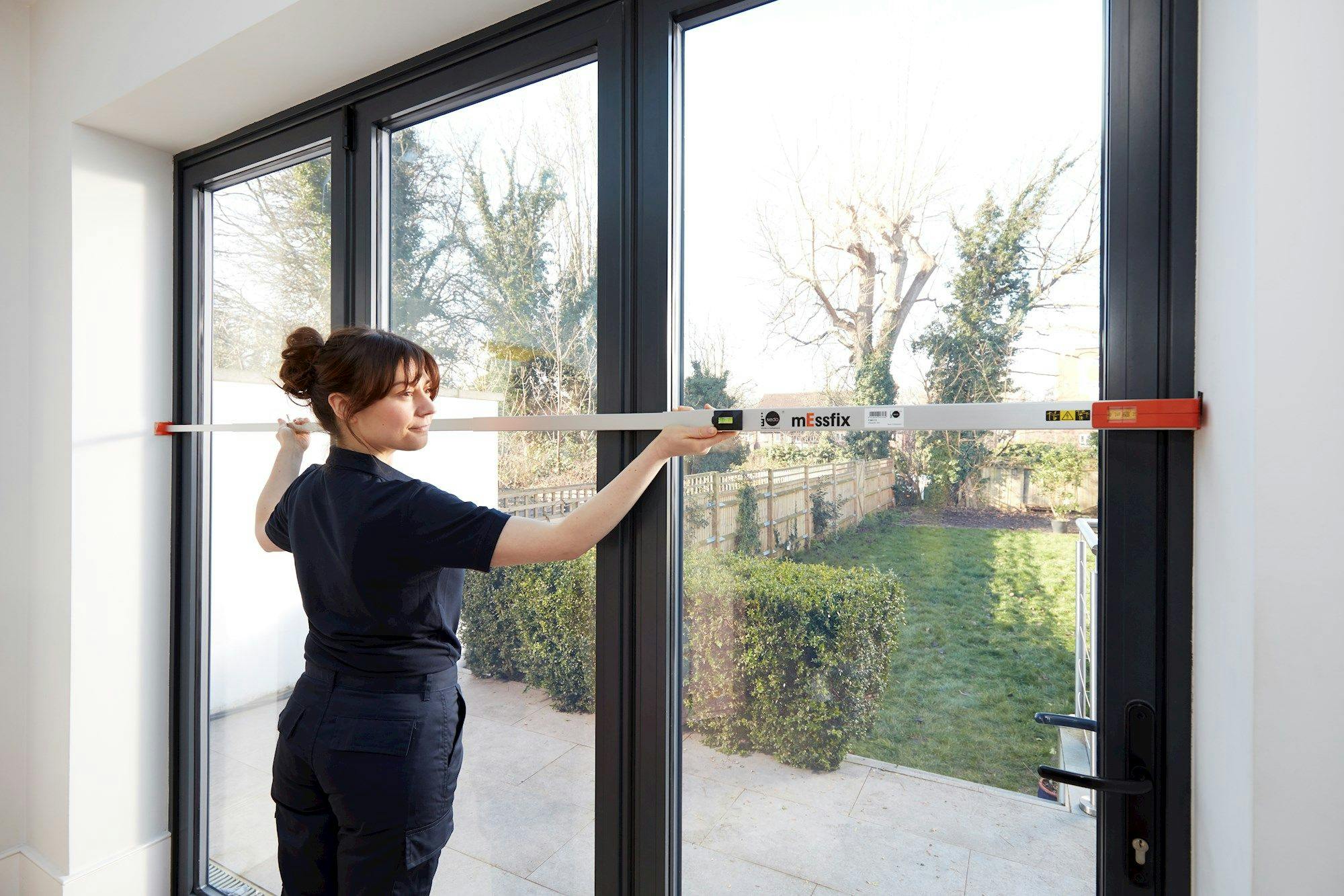 A person uses a measuring tool labeled "mEssfix" on a glass door, inside a room with an outside view of a garden with trees and a lawn.