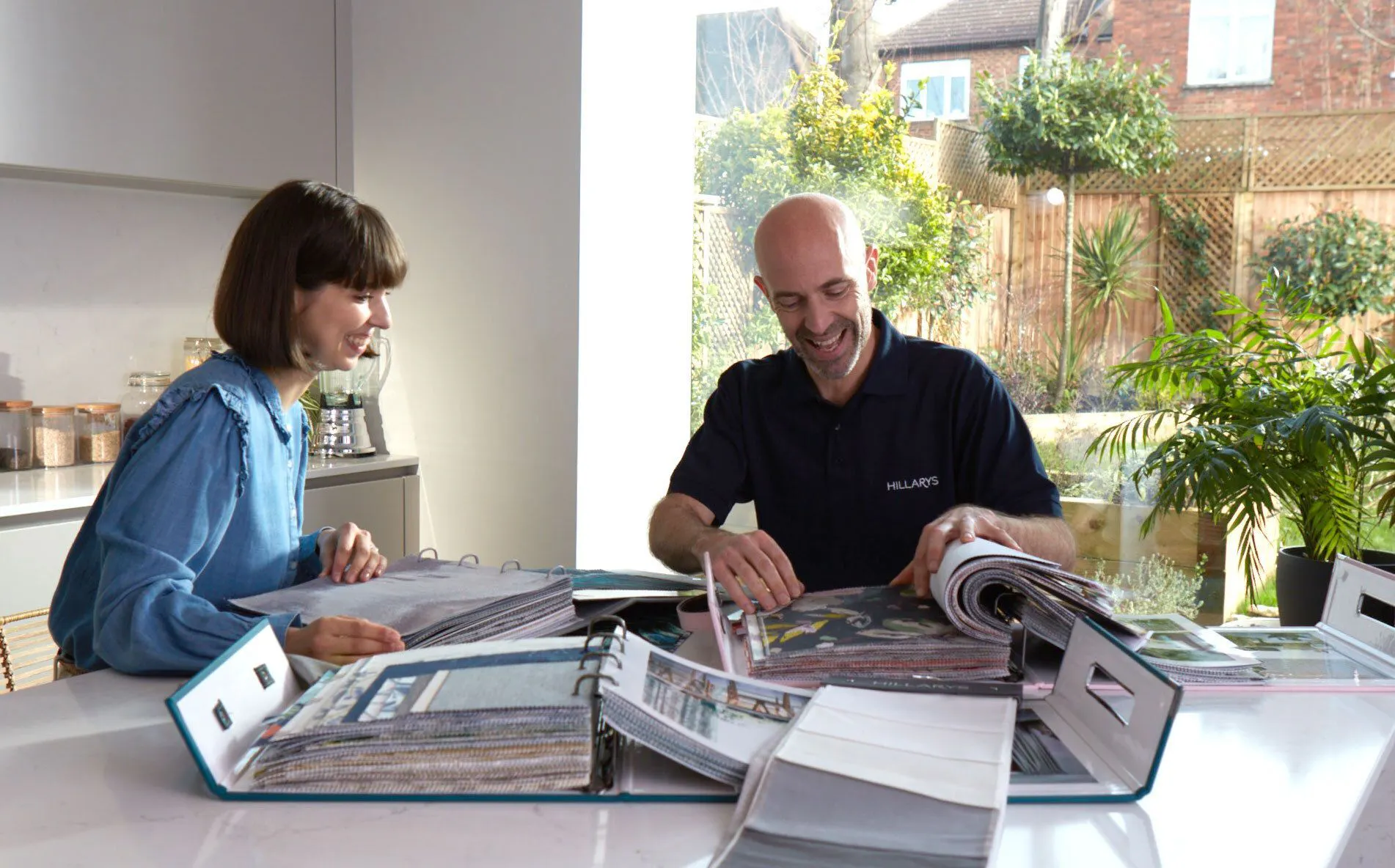 Two people sit at a table browsing through fabric sample books. One wears a "Hillary's" shirt. They're in a bright room with plants and a large window showing a garden.