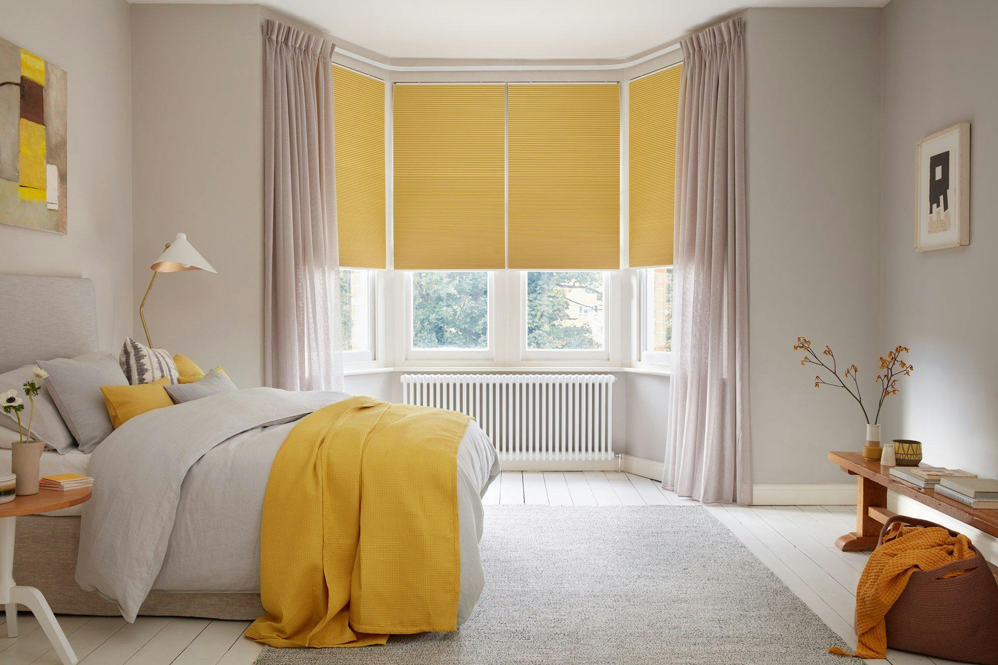 Bed draped in yellow blanket sits beside a lamp, beneath large windows with yellow blinds. Soft gray walls, light curtains, and a wooden table enhance the cozy bedroom atmosphere.