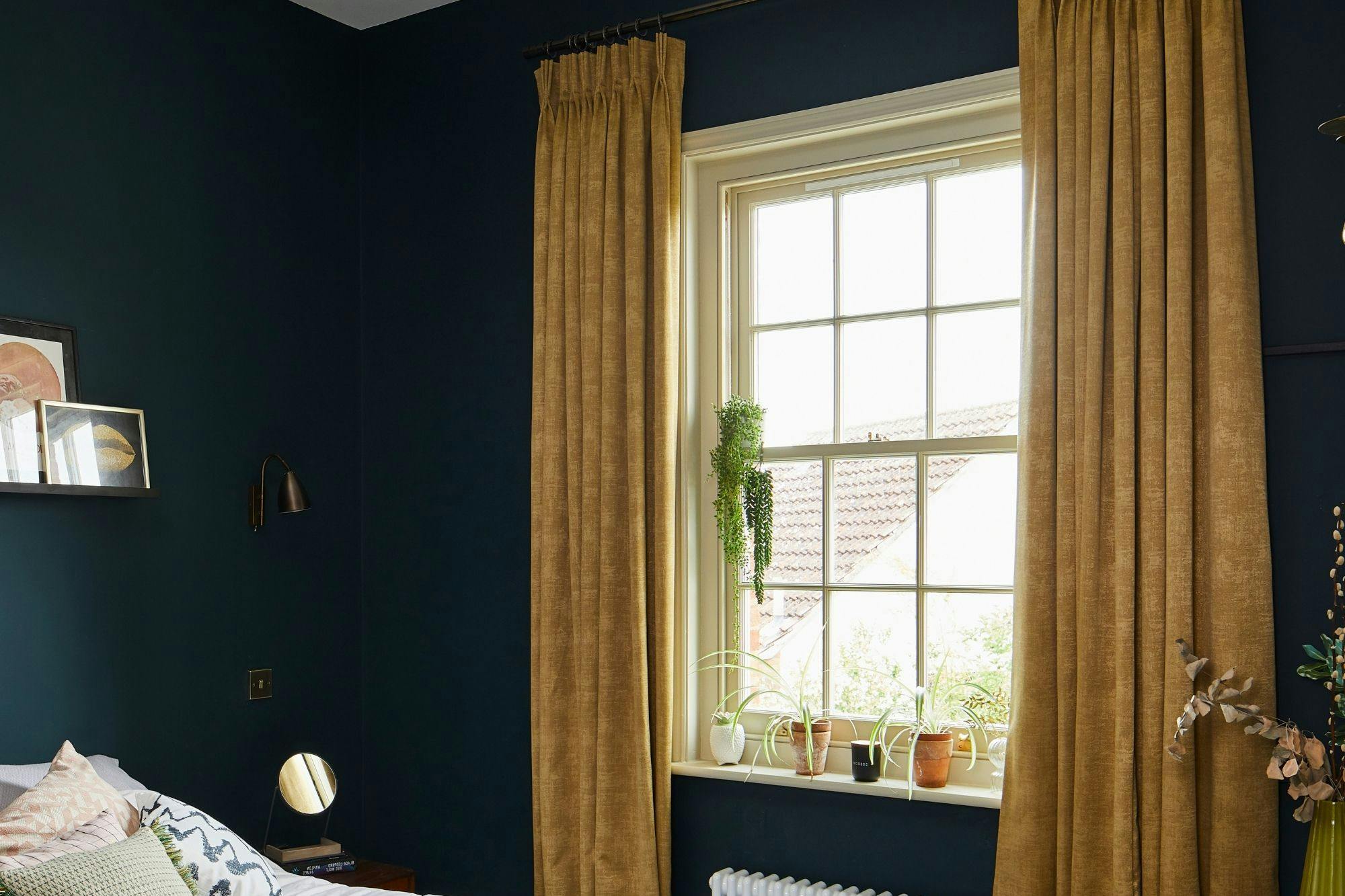 Mustard curtains in a bedroom. There is a bed on the left hand size that takes up the room. There is plotted plants in the room by the window and bed.