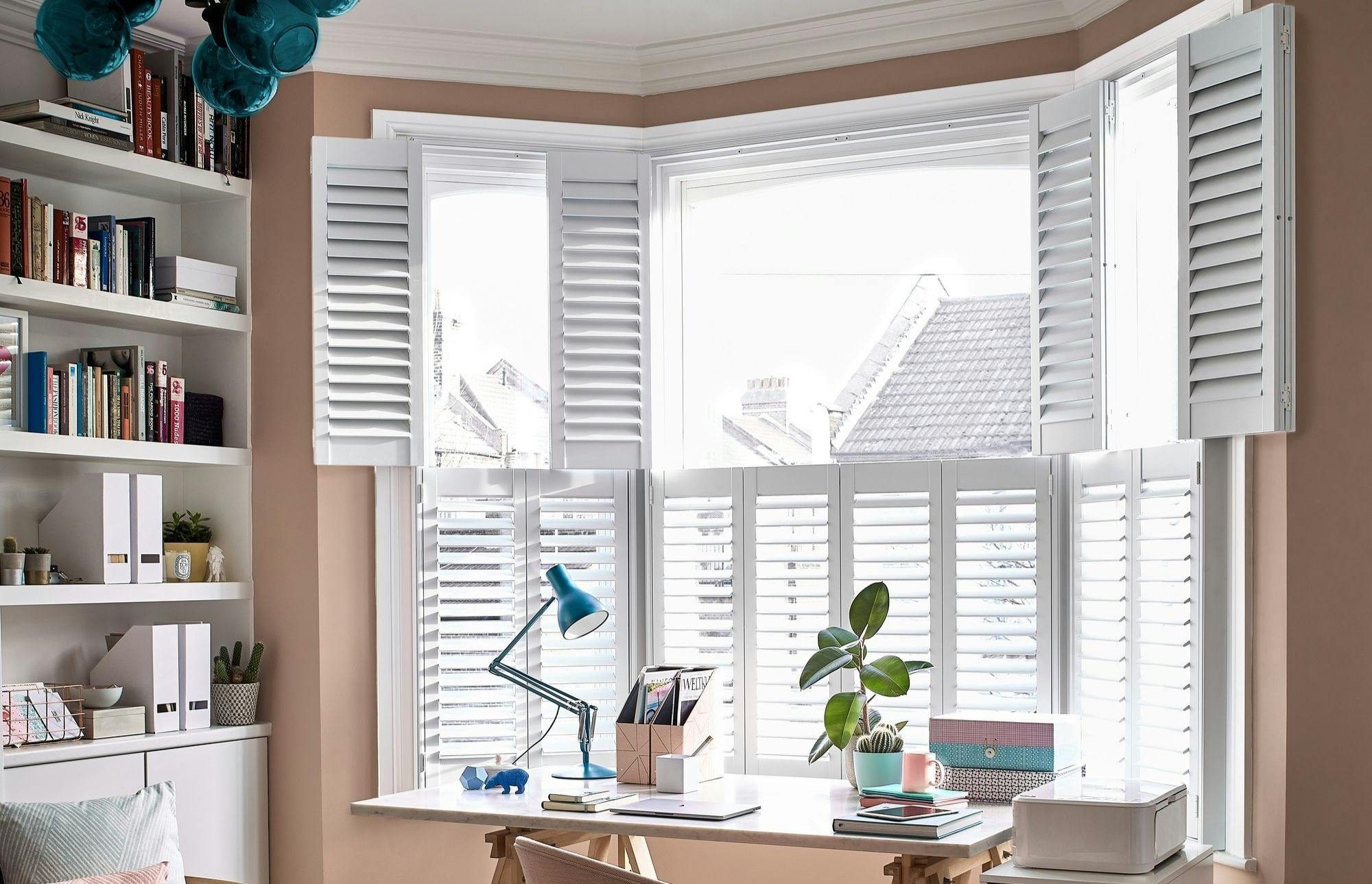 White tier on tier shutters in an office room. There is a desk with office equipment on it. There is a lamp and a potted plant on the desk. and a bookshelf on the left hand side of the room. 